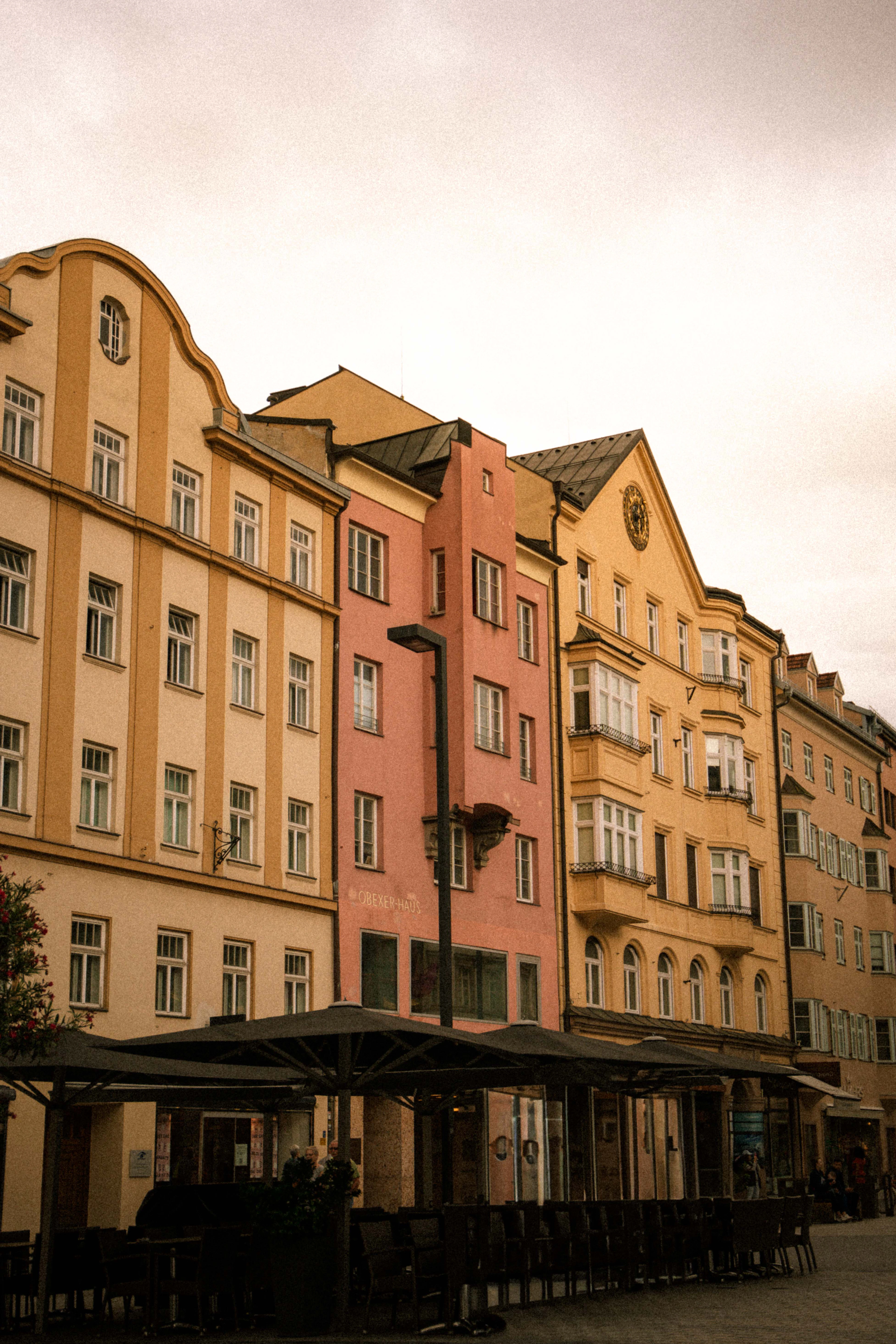 A row of tall Innsbruck townhouses in ochre, salmon pink and cream, with oriel windows, curved gables and wrought-iron balconies, standing above the furled parasols and empty chairs of a pavement cafe in warm evening light.