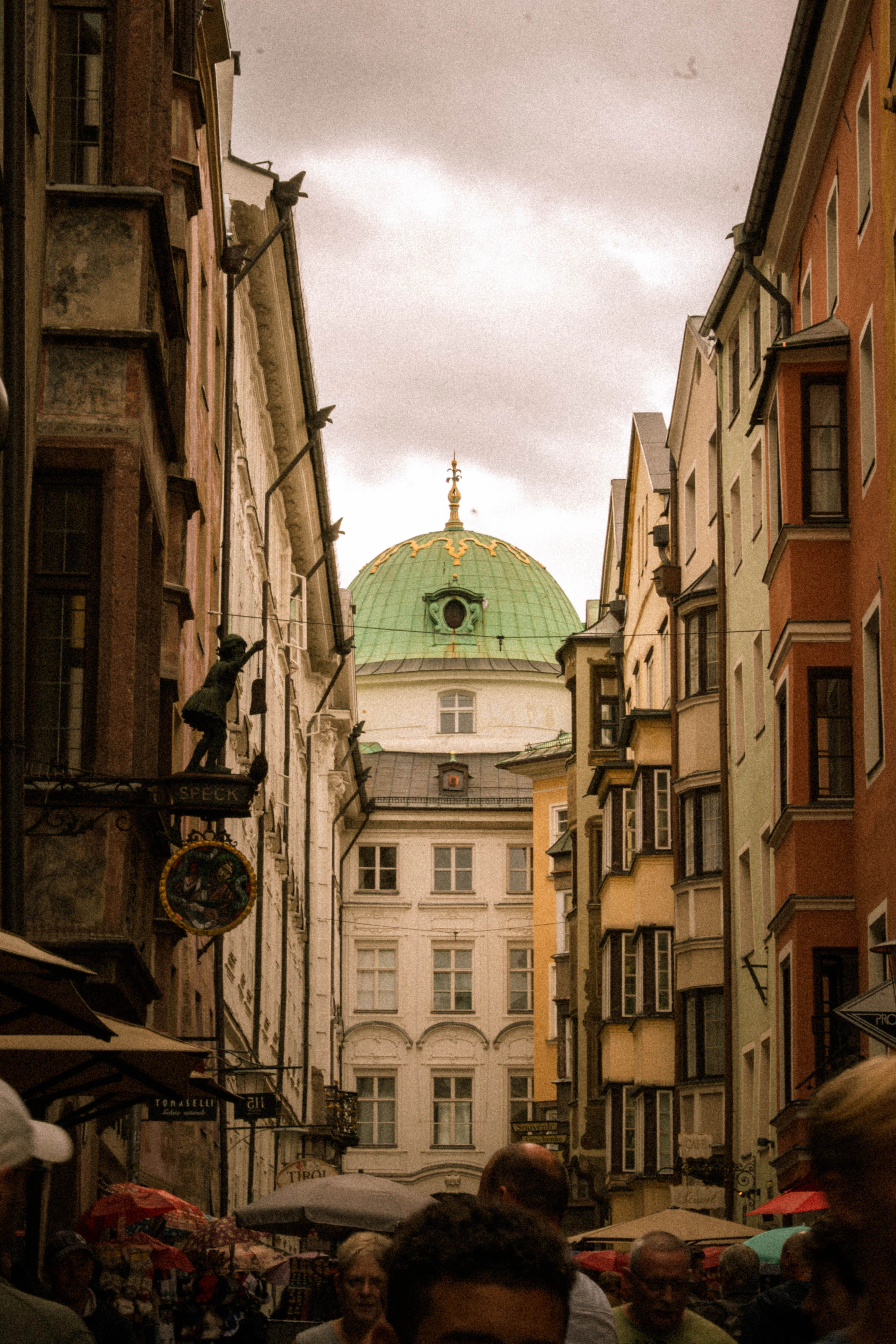 A narrow lane in Innsbruck's old town busy with summer visitors and cafe parasols, hand-painted wrought-iron shop signs projecting overhead and the green copper dome of a baroque church closing the view at the end.