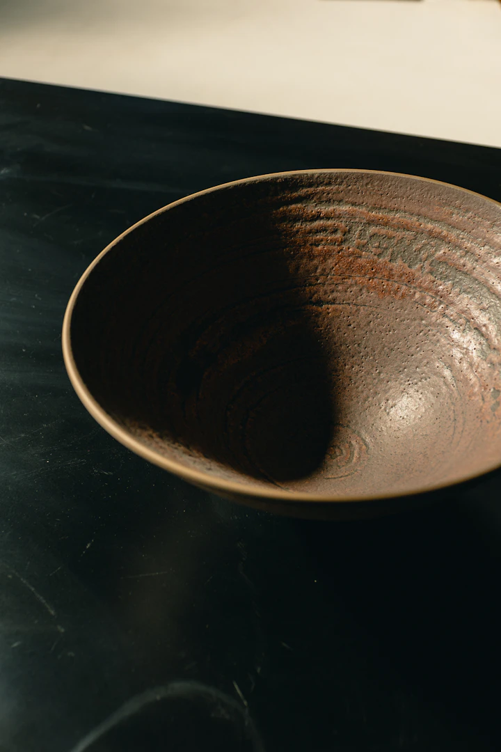 A single dark-glazed stoneware tea bowl on a black ground — Hamada-style chawan