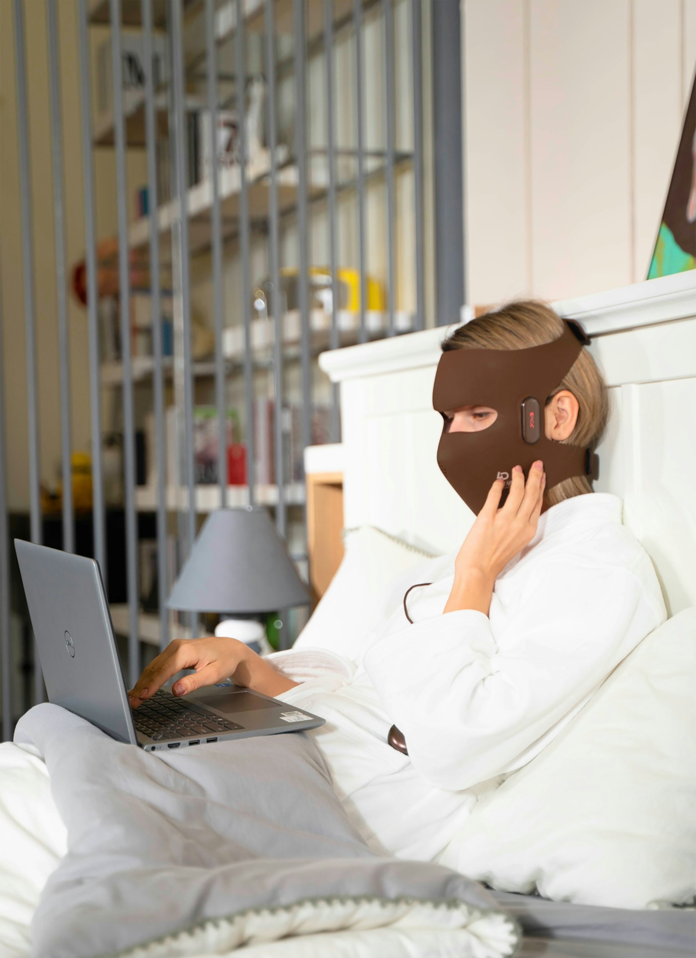 Woman relaxing in bed wearing an LED face mask while using her laptop