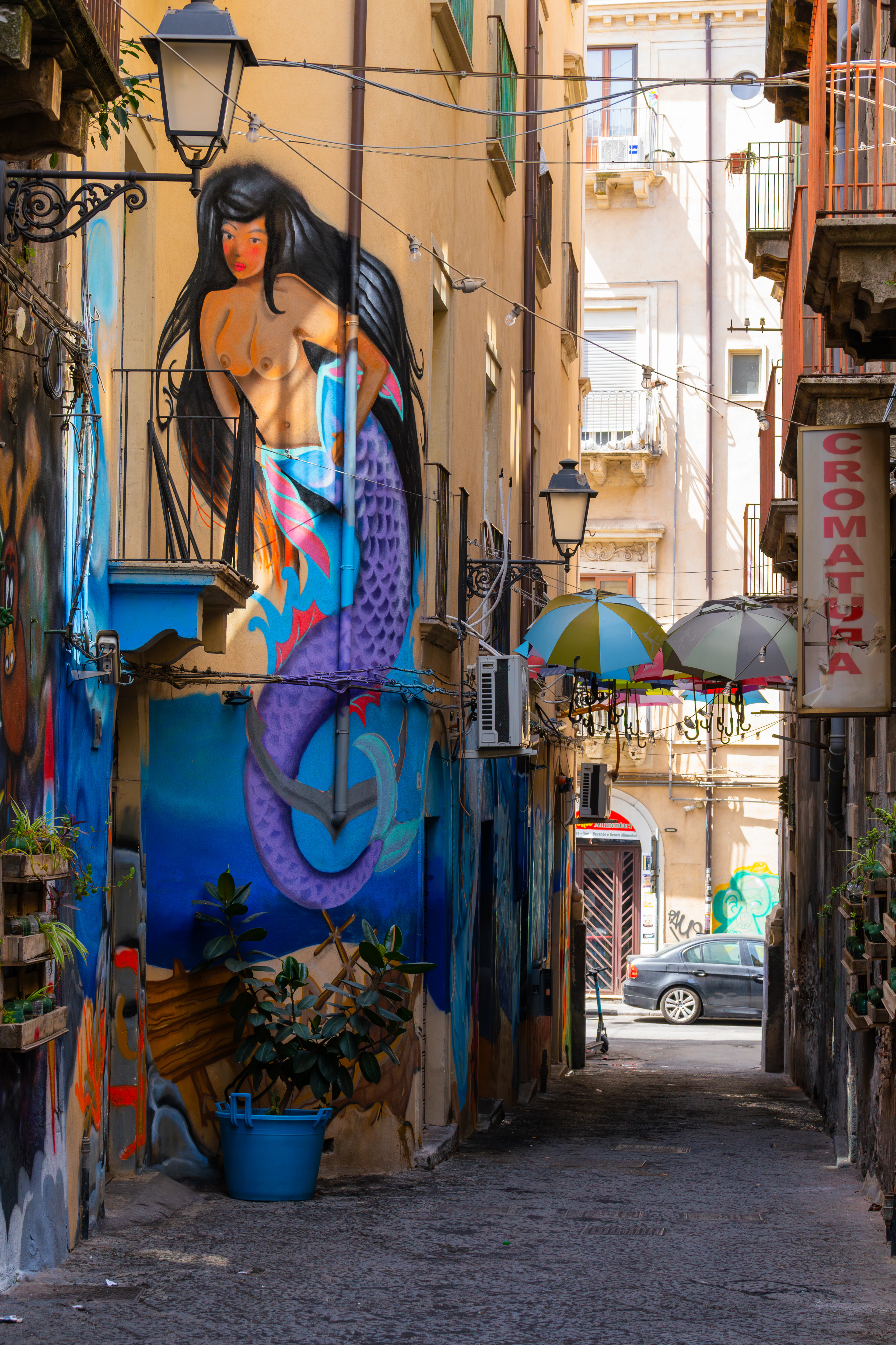 A narrow lane in central Catania with a large mermaid mural painted across the corner building, wrought-iron balconies and a hanging lantern above, and coloured cafe parasols catching the sun further down the street.