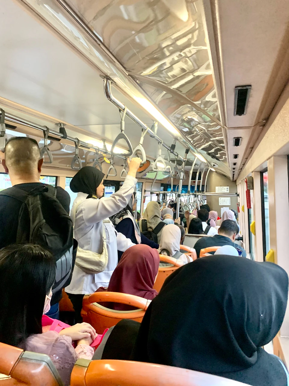 The image depicts a crowded subway car filled with people dressed in various colors, including black and white, pink, blue, and green. The passengers are seated on orange seats arranged in rows, facing forward towards the front of the train. Some individuals have their hands hanging over the open doors or handles, while others hold onto them for support as they stand up to exit the car.

The interior of the subway car is illuminated by a soft light that casts long shadows and highlights the textures of the seats and walls. The passengers appear engrossed in their own thoughts or conversations, creating an atmosphere of shared experience despite the crowded conditions.