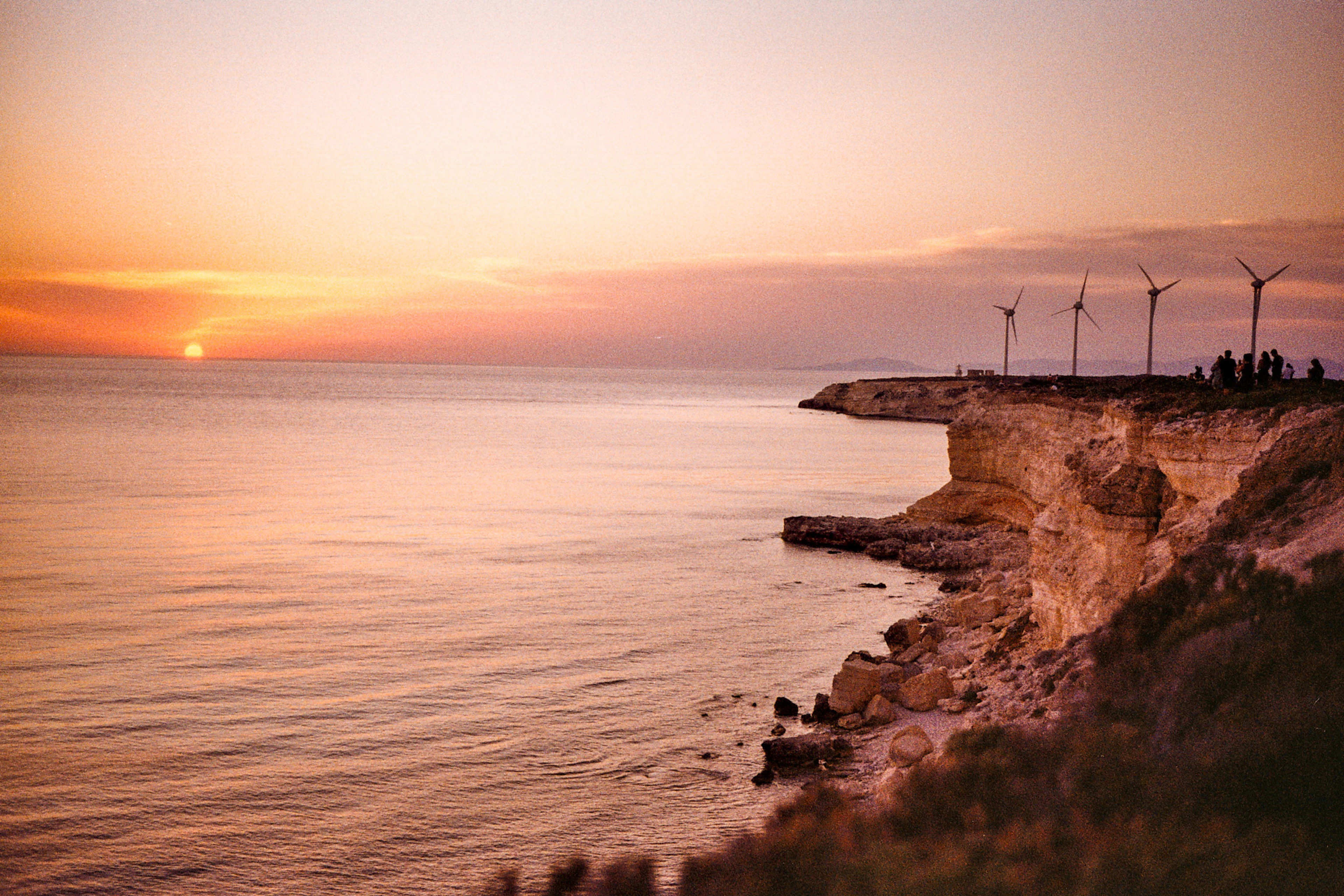 Sunset over the sea from a low rocky headland, with wind turbines and a small group of people on the cape