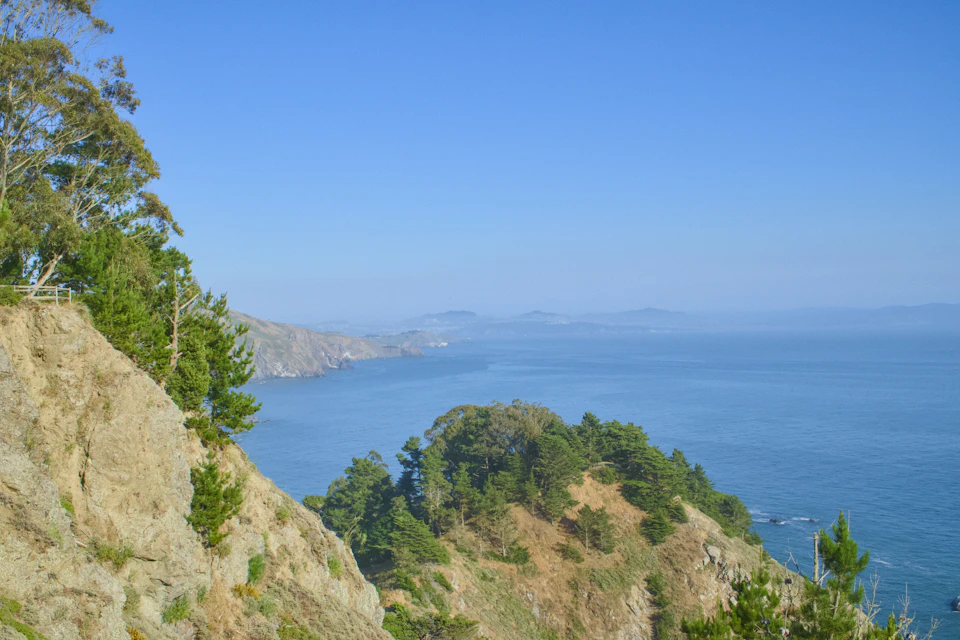 The image depicts a serene coastal landscape with a rocky cliff on the left side, overlooking a deep blue ocean that stretches out to meet the horizon. The sky above is a clear, bright blue color, and the water below mirrors its hue in a darker shade of blue. On the right side of the image, there are trees scattered across the scene, adding a touch of green to the otherwise rocky landscape.