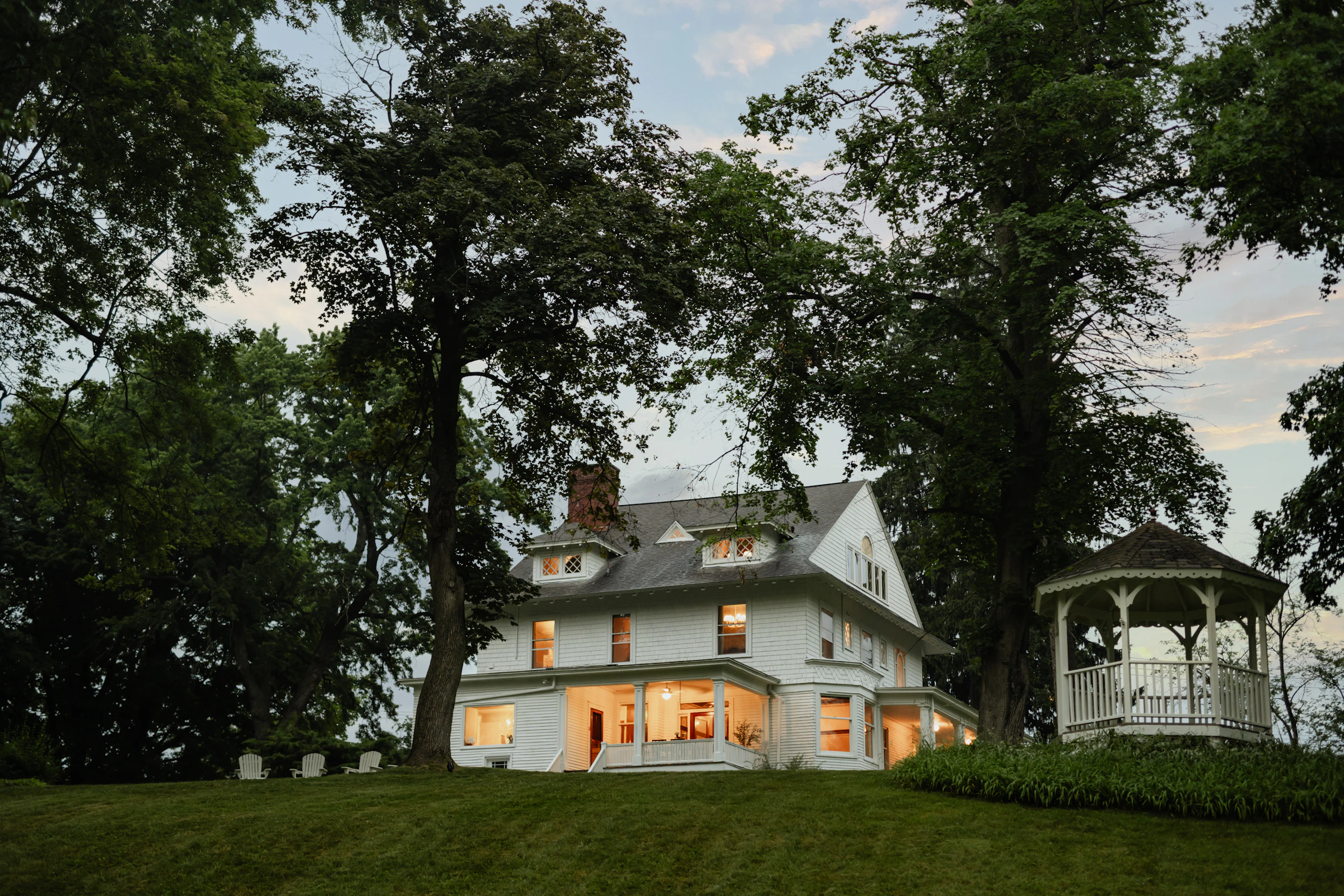 A white two-story house glowing at dusk among old shade trees, with a gazebo beside it