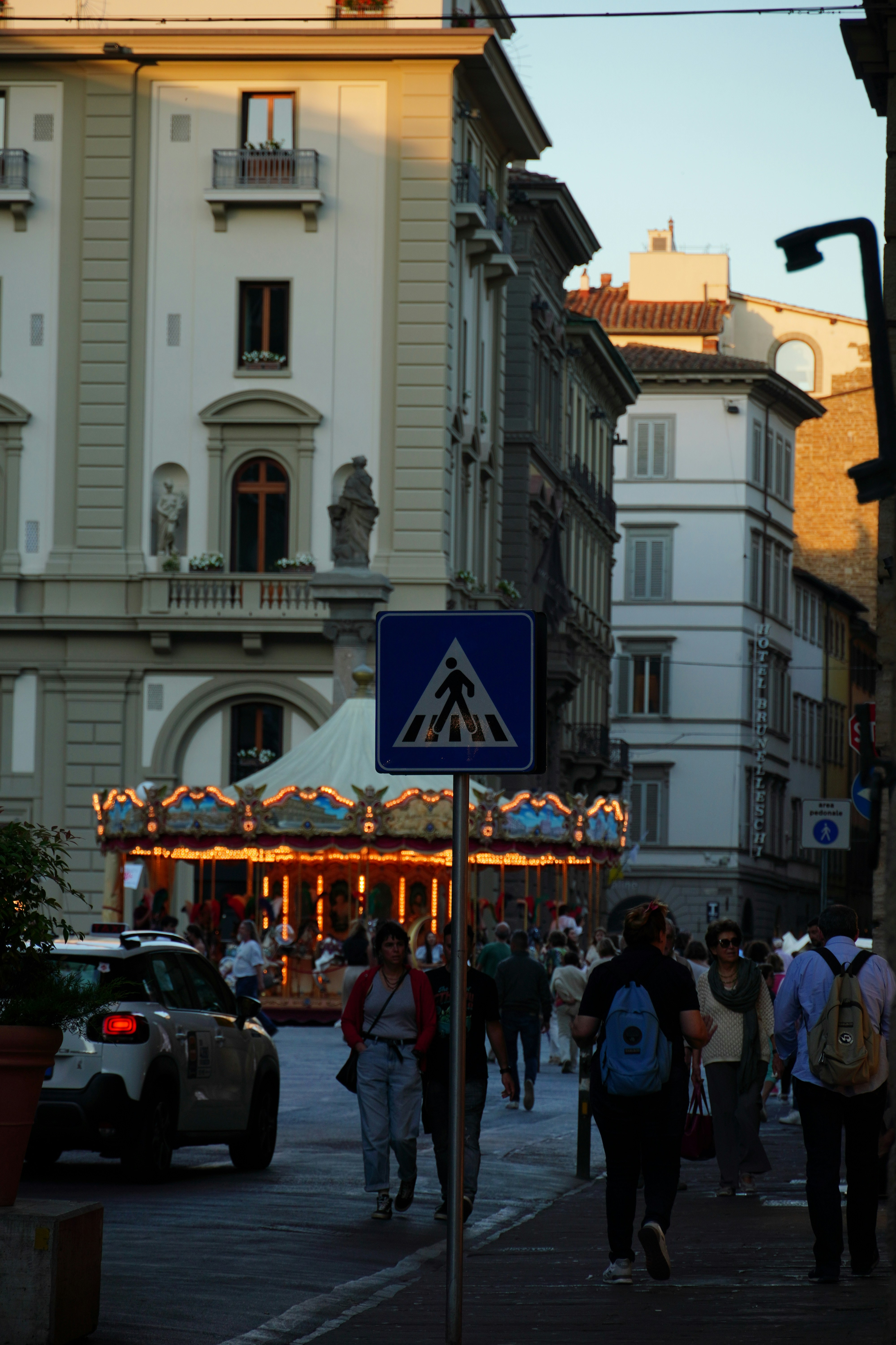 A lively city street with people walking, a pedestrian sign, and a carousel in the background at dusk.