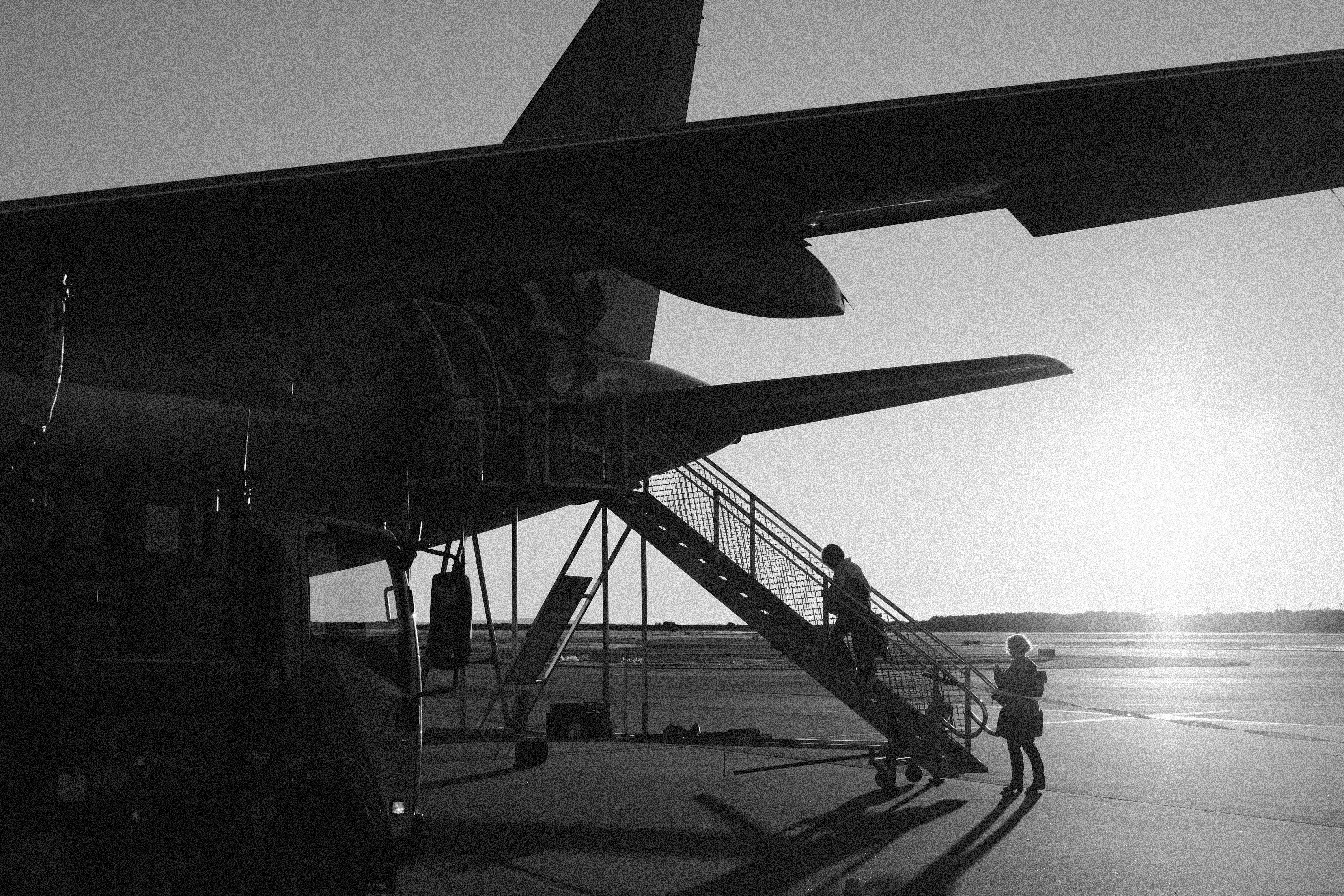 A large jetliner sitting on top of an airport tarmac, A black and white image capturing the silhouette of an Airbus A320 as passengers board the aircraft at sunrise. The photo highlights the serene moment of morning light casting long shadows across the tarmac. The scene features a person standing at the base of the boarding stairs and another person ascending the stairs, adding a sense of journey and anticipation. The minimalistic composition and high contrast create a timeless and evocative atmosphere.