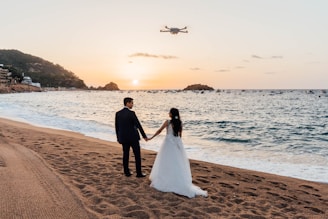 A bride and groom walking on the beach at sunset