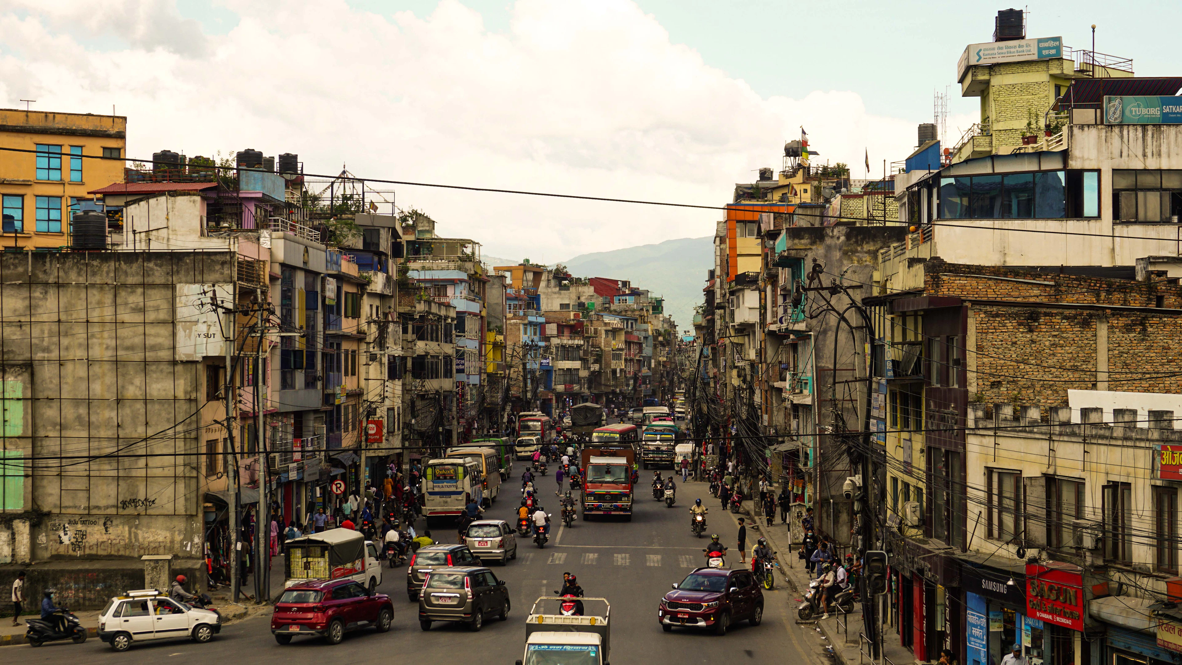 A busy Kathmandu avenue seen from above, packed with buses, taxis, motorbikes and an auto-rickshaw between tall painted shophouses, with the green valley rim hazy in the distance