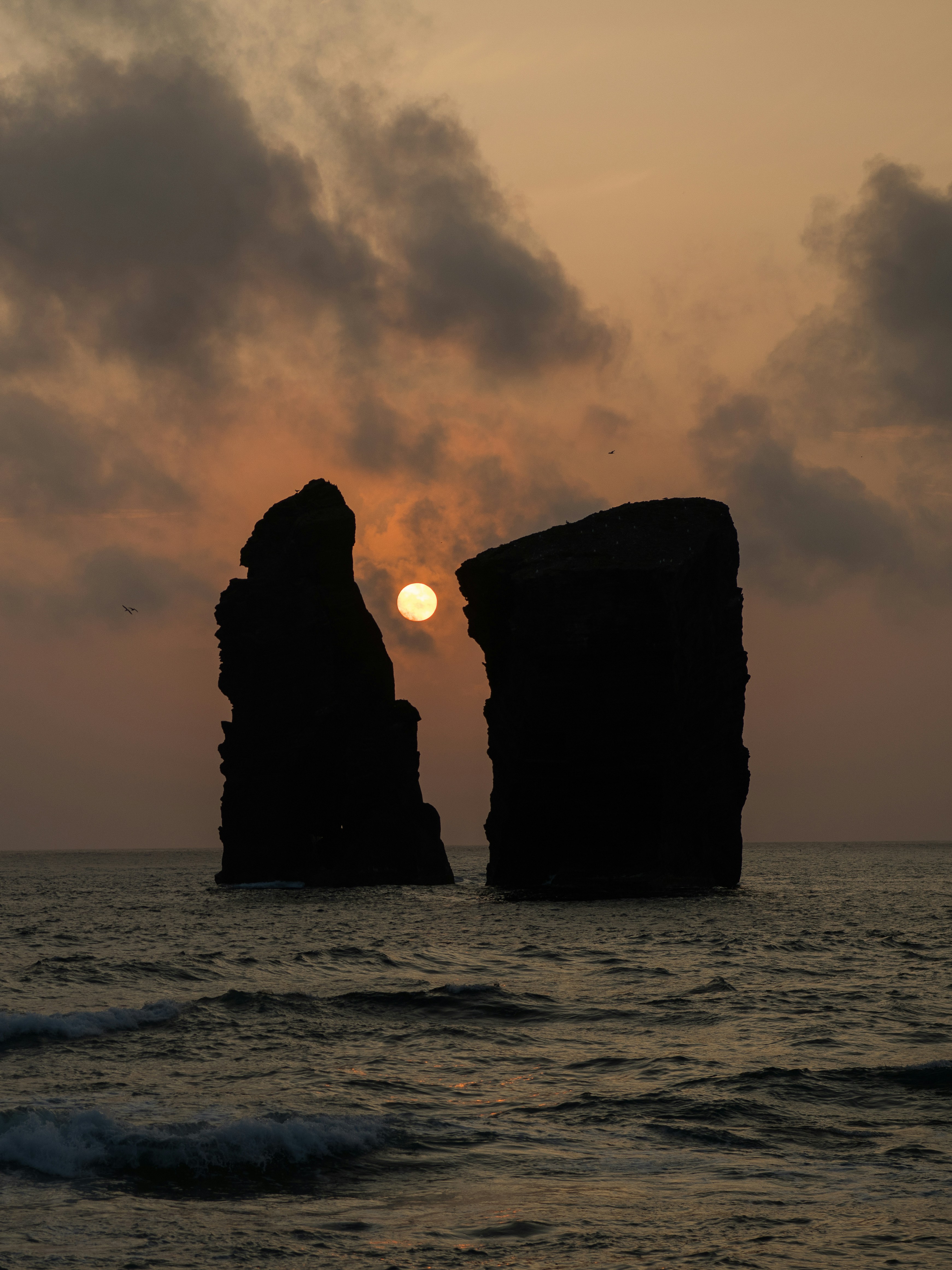 The sun is setting behind two large rocks in the ocean