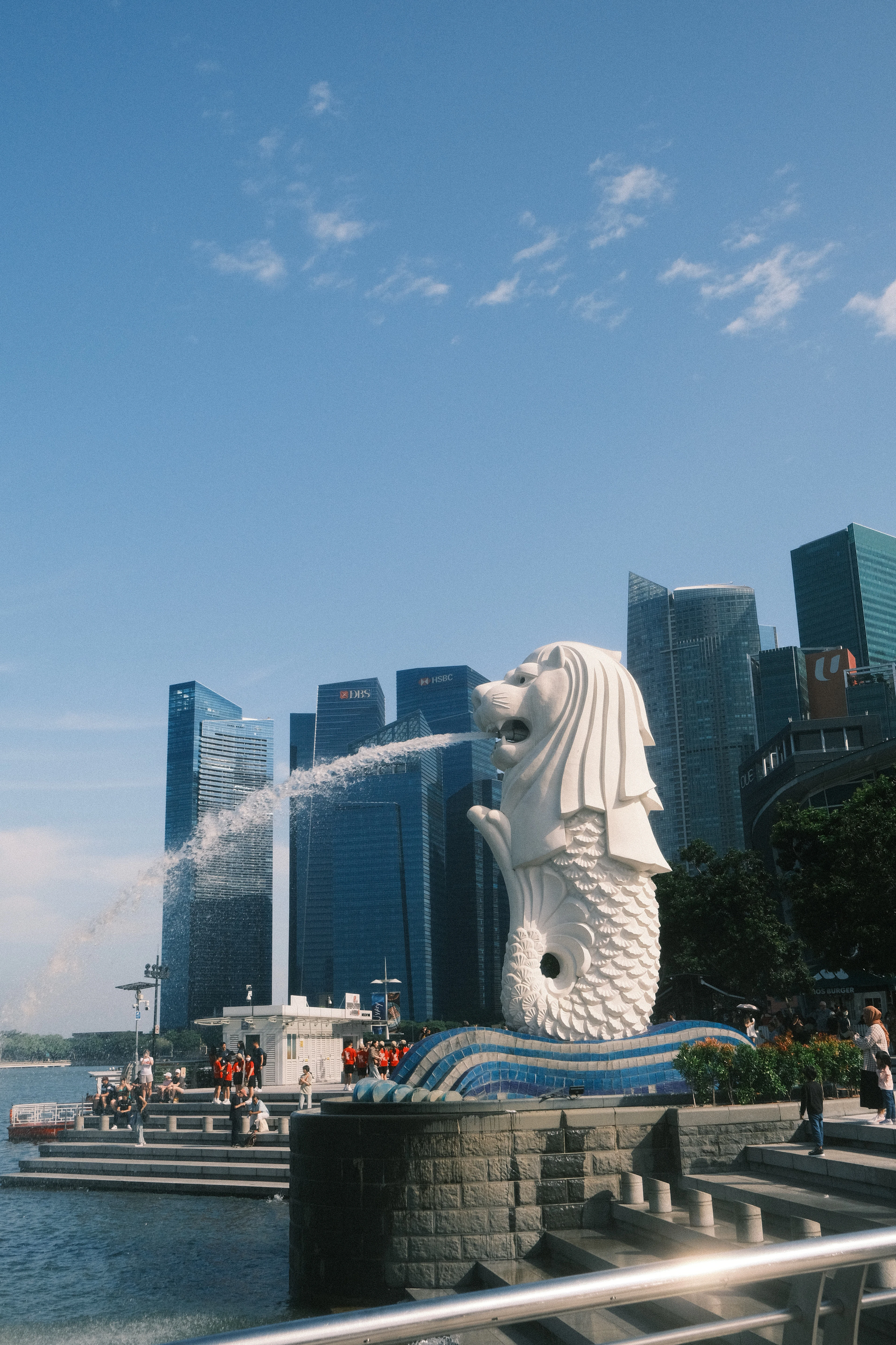 The Merlion fountain sprays water into Marina Bay against a clear blue sky and the downtown skyline.