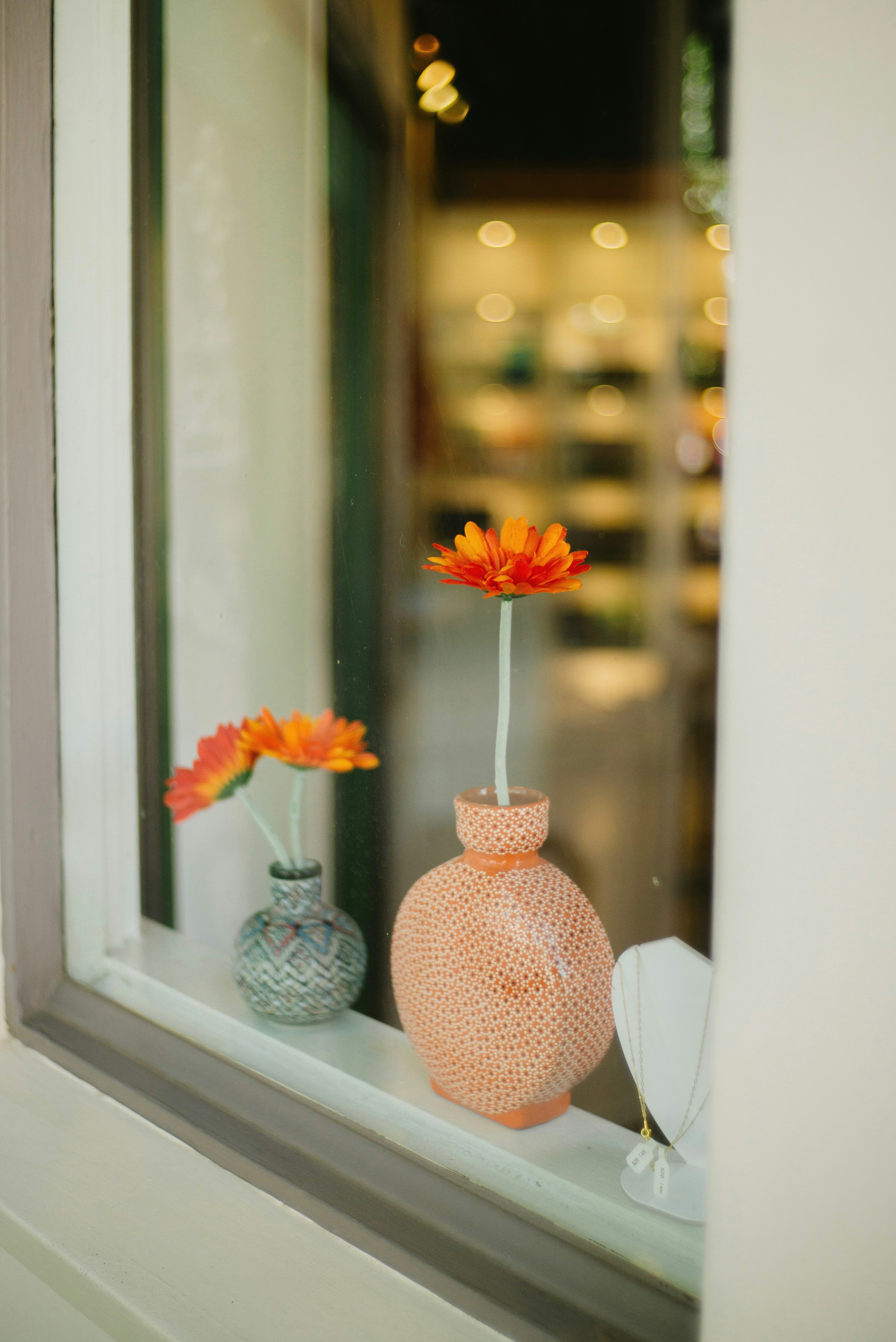 Colorful orange flower in a textured ceramic vase sits on a windowsill with blurred shop lights in the background.