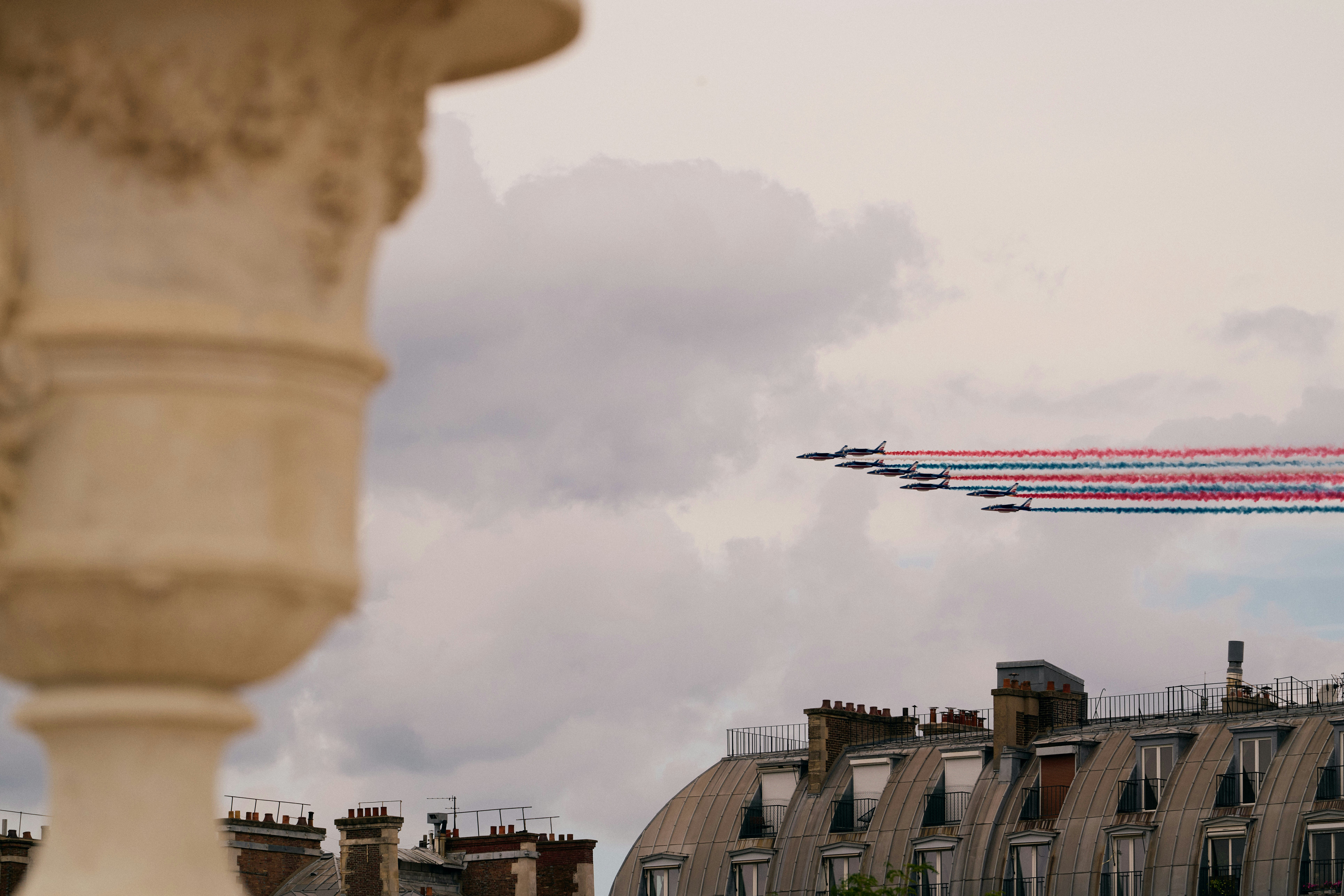 French Air Force Aerobatic Display Over Paris Description: A striking photograph capturing a formation of French Air Force jets trailing red, white, and blue smoke as they fly over the rooftops of Paris. The jets are in perfect alignment, showcasing their precision and skill against a cloudy sky. In the foreground, the out-of-focus architectural details add a sense of depth and contrast to the dynamic scene above. This image beautifully encapsulates the blend of historic and contemporary elements that define Parisian charm and national pride. Ideal for themes of aviation, patriotism, and urban landscapes.