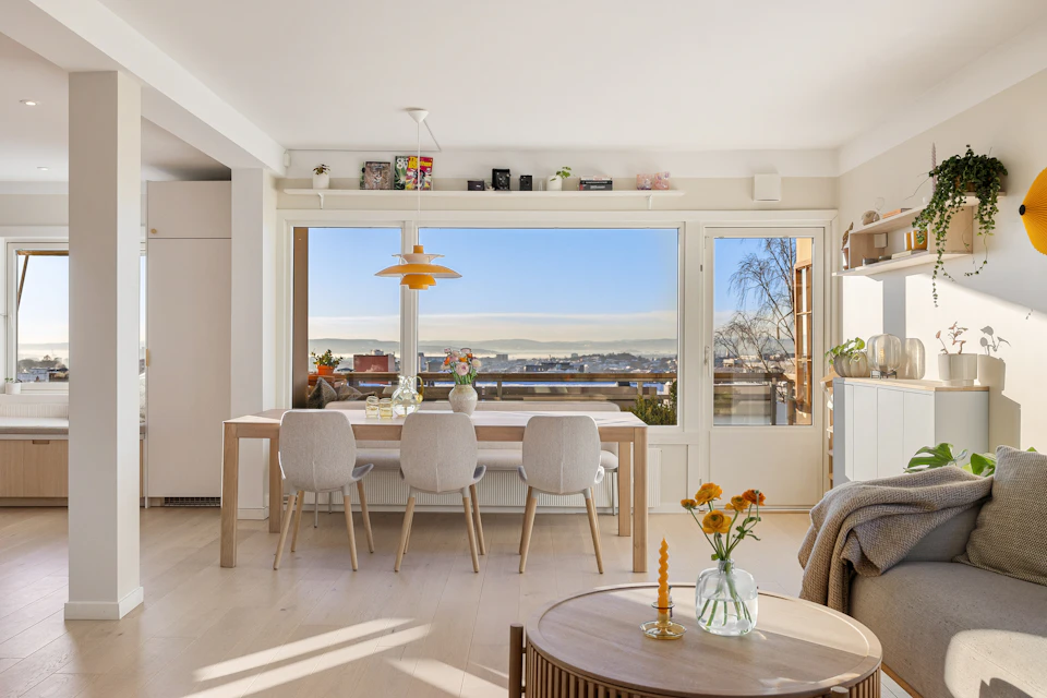 The image depicts a modern living room with a large window that offers a view of a cityscape, allowing natural light to flood the space. The room is furnished with white furniture and features a wooden coffee table adorned with a vase filled with flowers in shades of orange and yellow. A gray sofa sits against one wall, while another chair is positioned near the window. A bookshelf stands tall on the opposite side of the room, housing an array of books and decorative items. The walls are painted white, creating a bright and airy atmosphere, and the floor is made of wood, adding warmth to the space.
