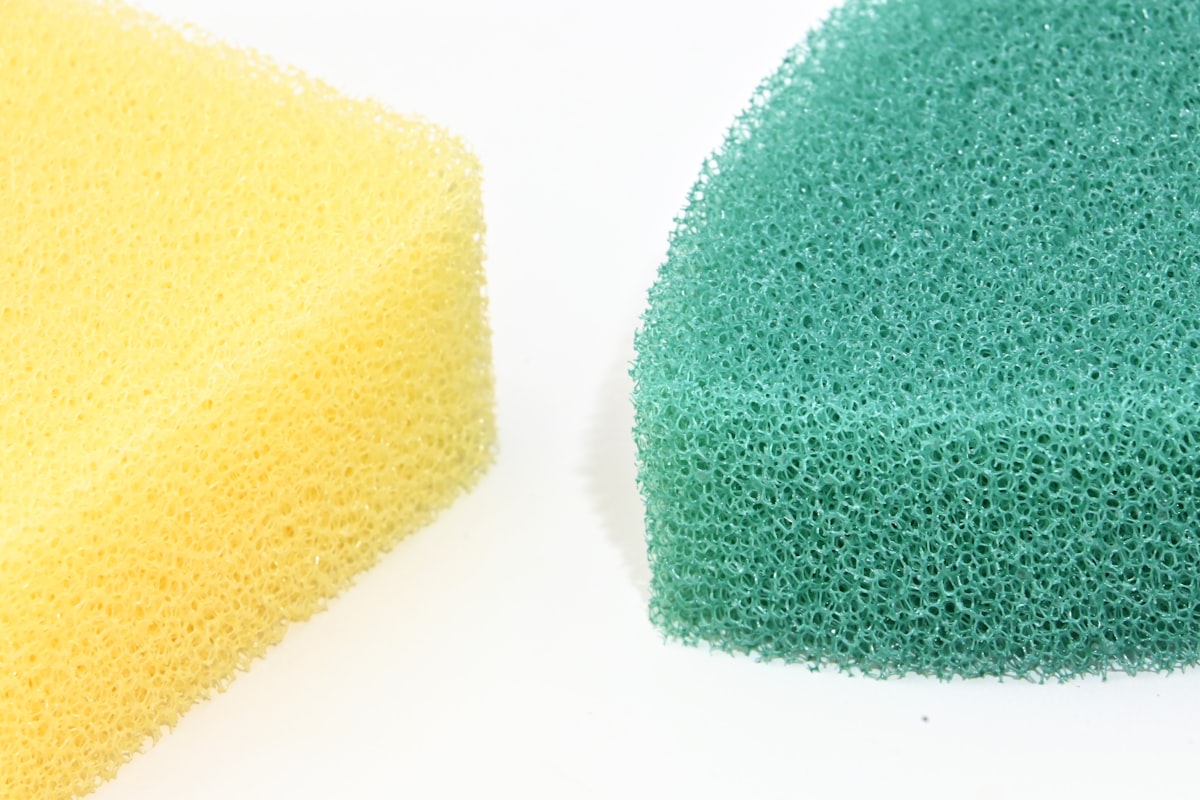 Dos esponjas de cocina en primer plano sobre fondo blanco, una amarilla y una verde, mostrando su textura porosa llena de poros