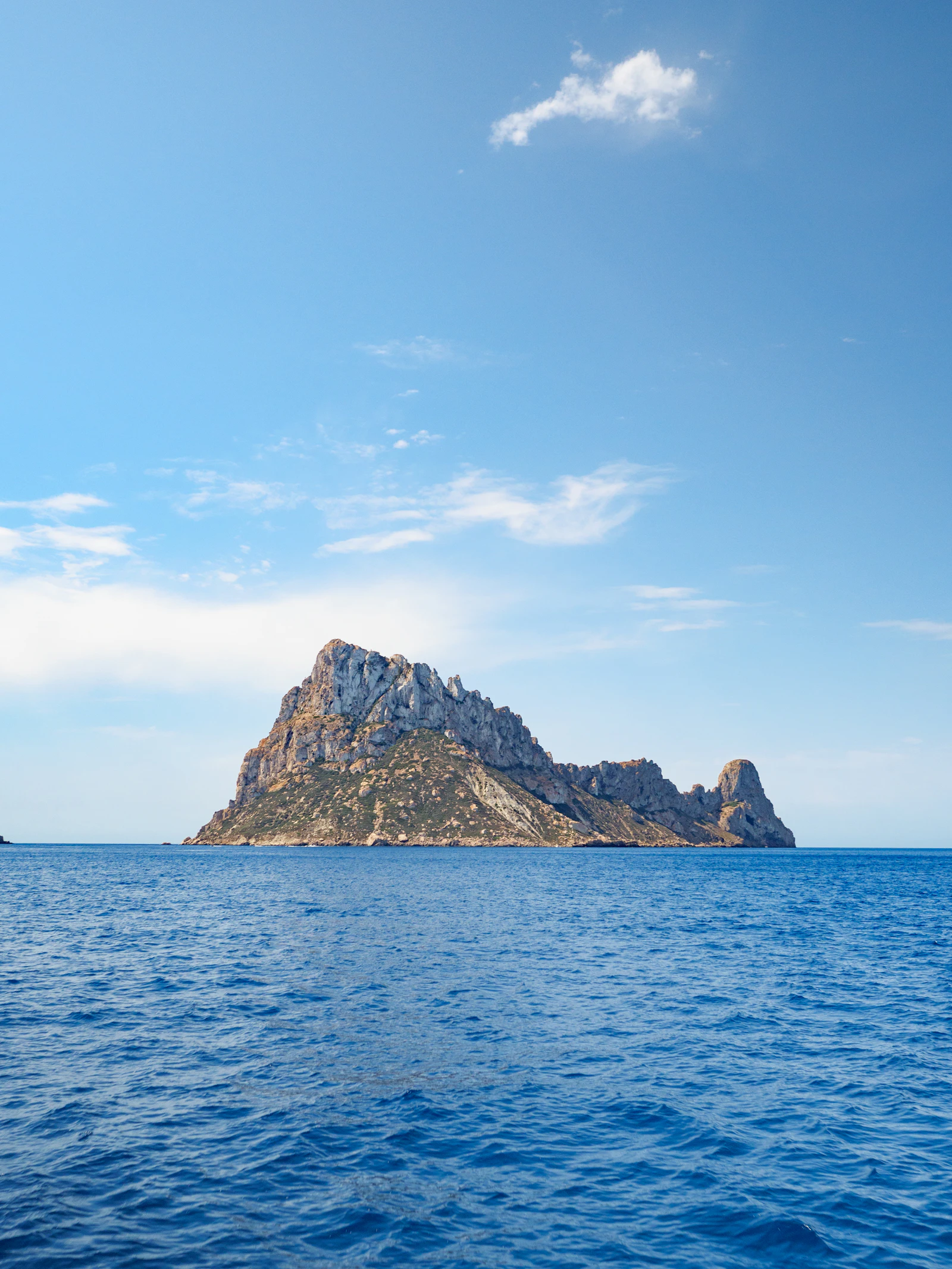 Es Vedrà rising from the blue Mediterranean off Ibiza's rocky coastline beneath a bright summer sky