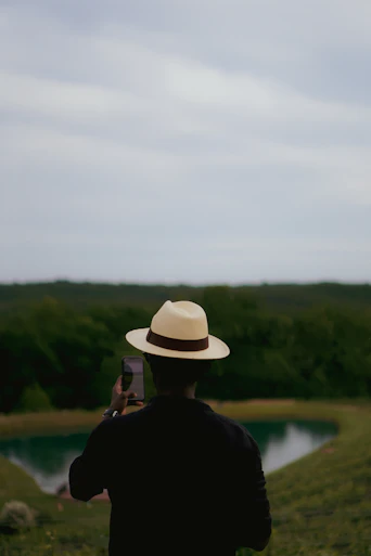 A man in a hat taking a picture of a lake