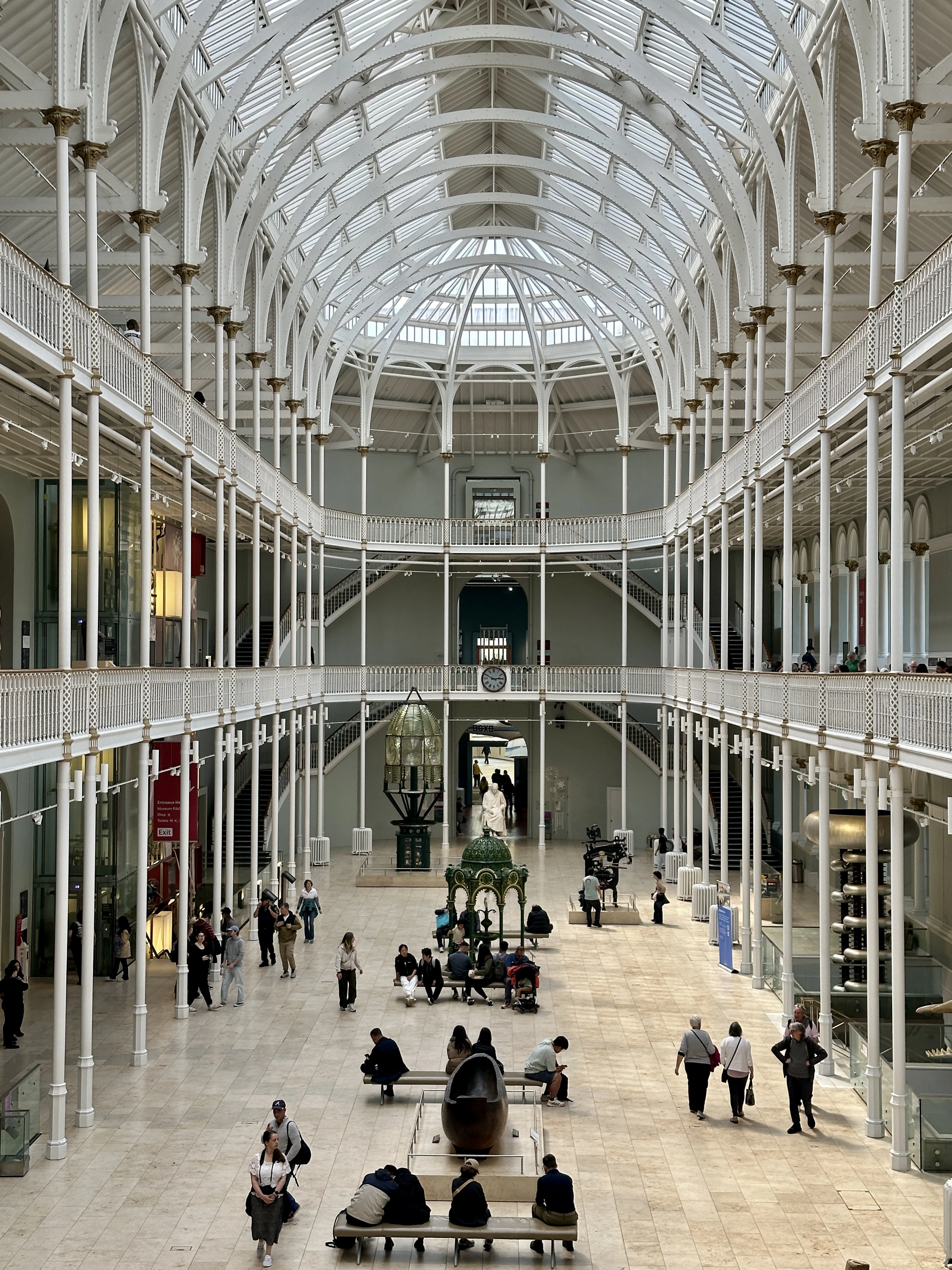 The Grand Gallery of the National Museum of Scotland in Edinburgh, white cast-iron arcades rising through two balconied tiers to a barrel-vaulted glass roof and domed skylight above a pale sandstone floor where visitors sit and wander.