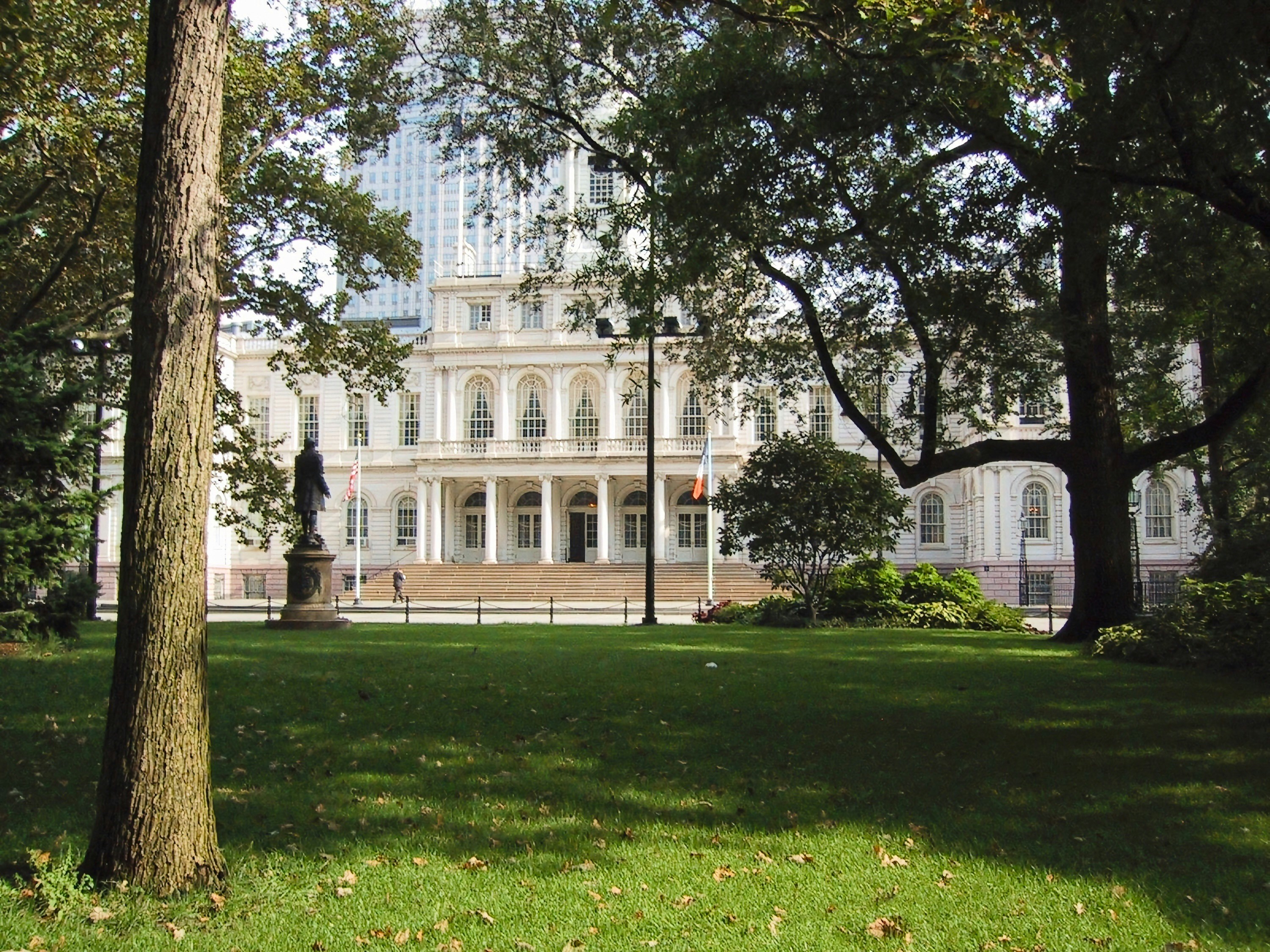 A Neo-Classical building façade with tall columns rises behind a manicured lawn, framed by trees and a statue on the left.