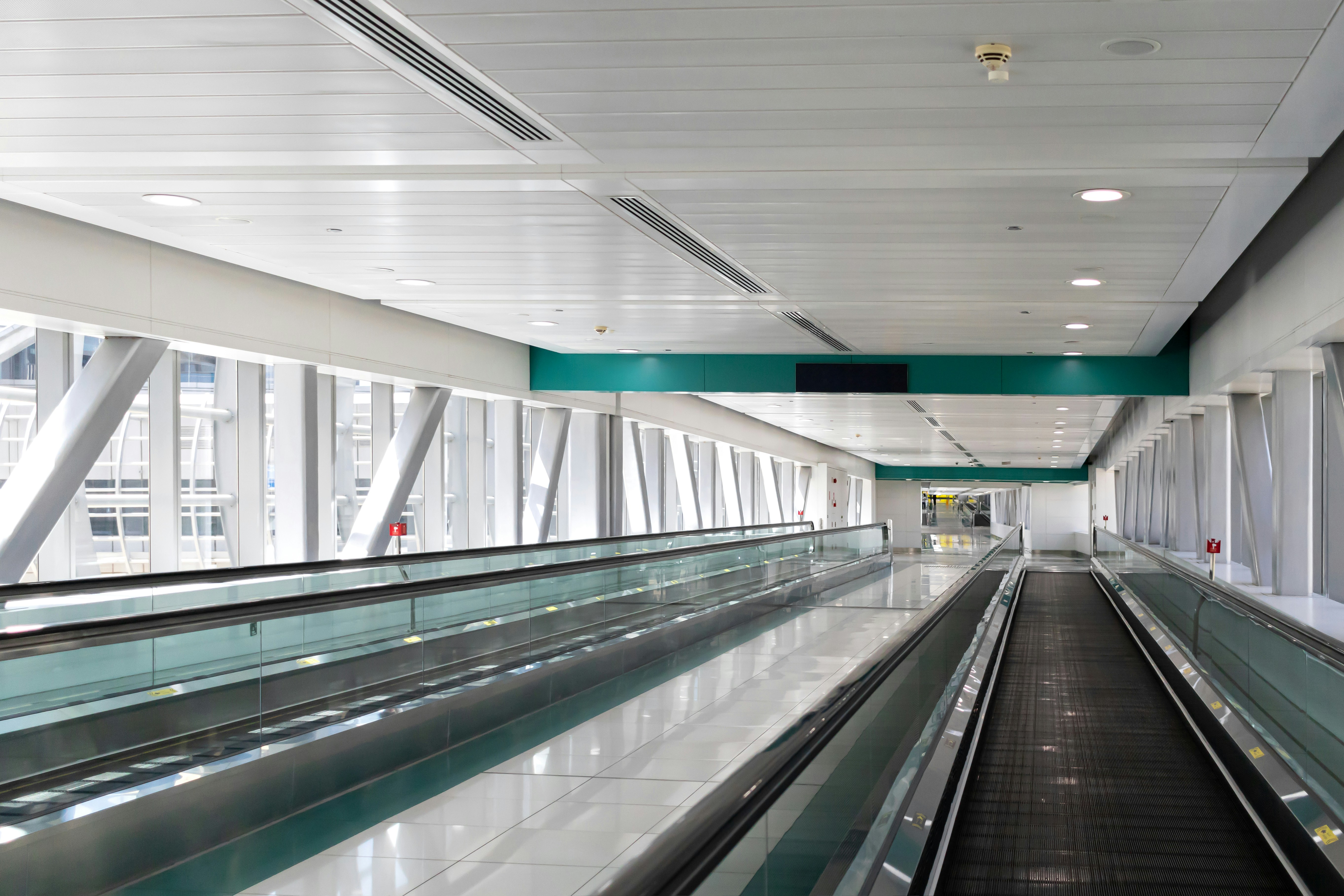 A view of an empty subway station from the bottom of the escalator, A long modern transit hallway with a green and white ceiling and a green stripe on the ceiling