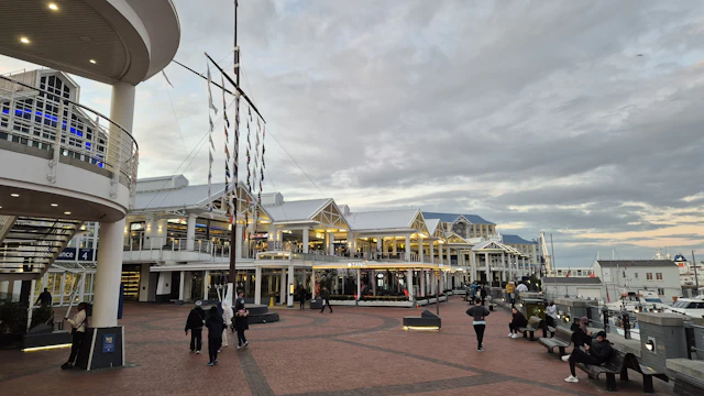 The V&A Waterfront promenade lined with Victorian-era warehouse buildings and a ship's mast sculpture, people walking along the quay.