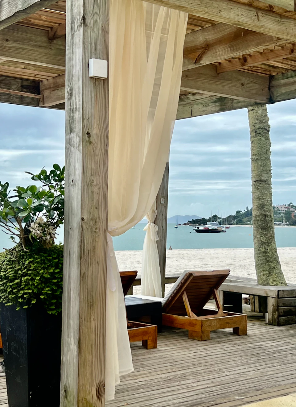 Two wooden lounge chairs sit under a large window with beige curtain on patio
