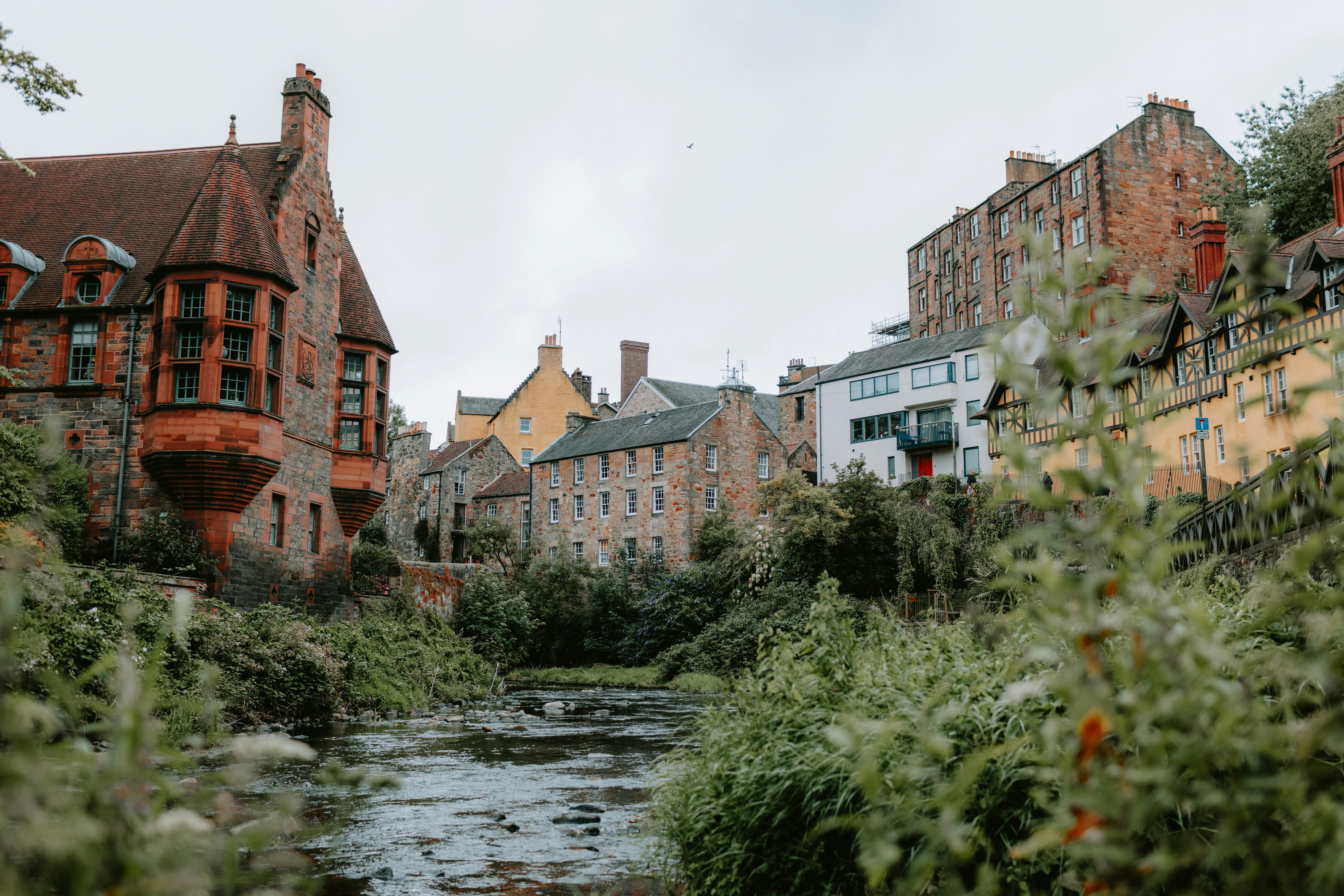 A river running through a lush green forest next to tall buildings, 