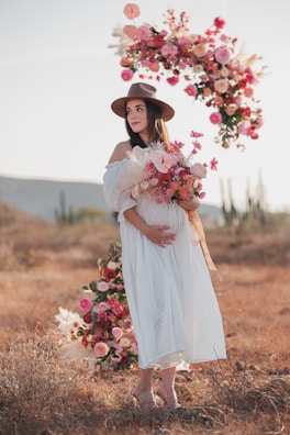 A pregnant woman in a white dress and hat holding a bouquet of flowers