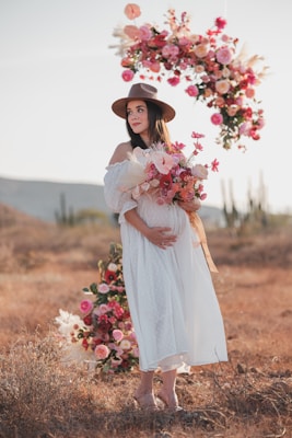 A pregnant woman in a white dress and hat holding a bouquet of flowers
