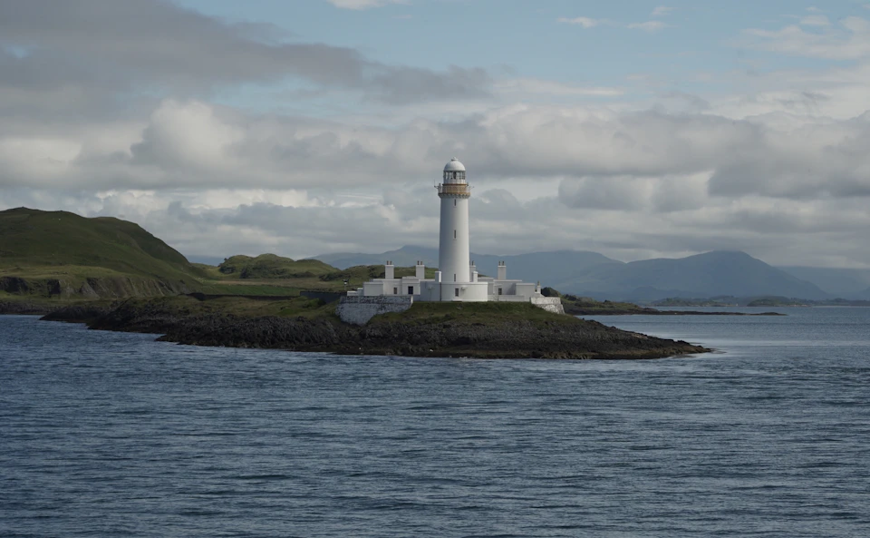 The image depicts a serene scene at sea, featuring a large white lighthouse standing tall on an island amidst the vast expanse of water. The lighthouse is surrounded by a rocky shoreline and a grassy field, adding to its picturesque charm. In the distance, mountains can be seen rising against the horizon, providing a stunning backdrop for this tranquil setting.