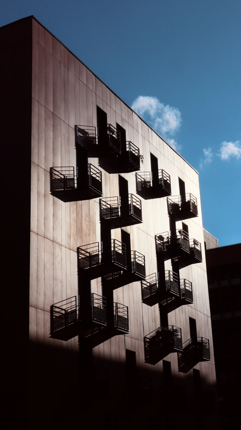 Tall rectangular building with concrete and metal facade, multiple balconies with black railings visible