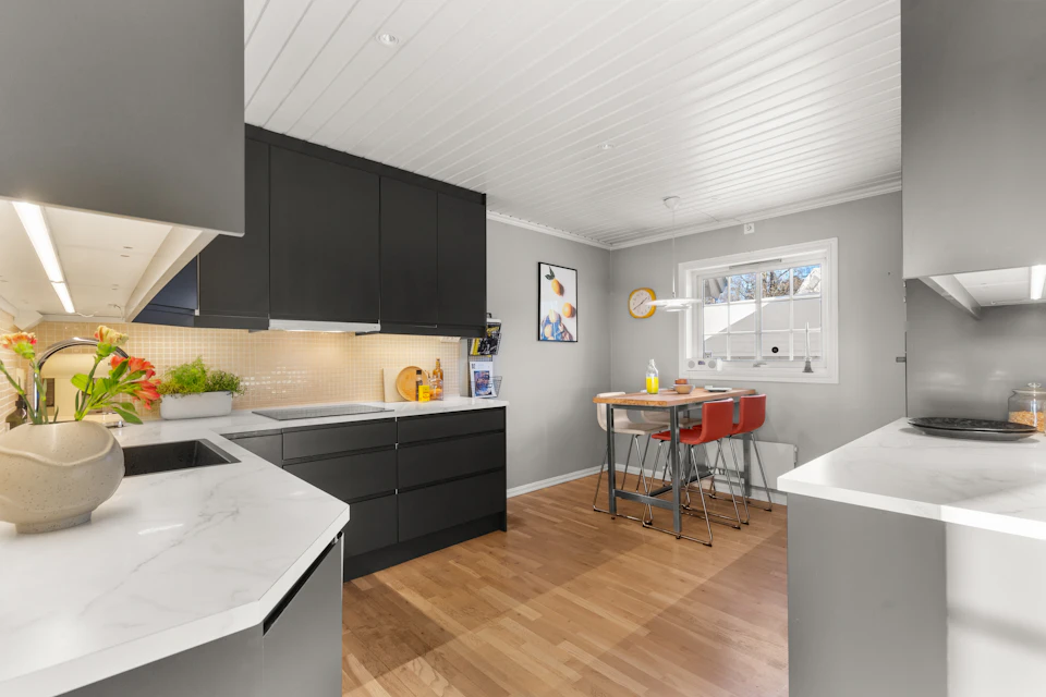 The image depicts a modern kitchen with white walls and gray cabinets, creating a clean and minimalist aesthetic. The kitchen features black countertops that contrast with the white surroundings. A window above the sink allows natural light to enter the space, enhancing its brightness. On one side of the room, there is a dining table surrounded by four chairs, suggesting an area for socializing or enjoying meals together. In front of the dining table, there are two potted plants adding a touch of greenery and life to the space.