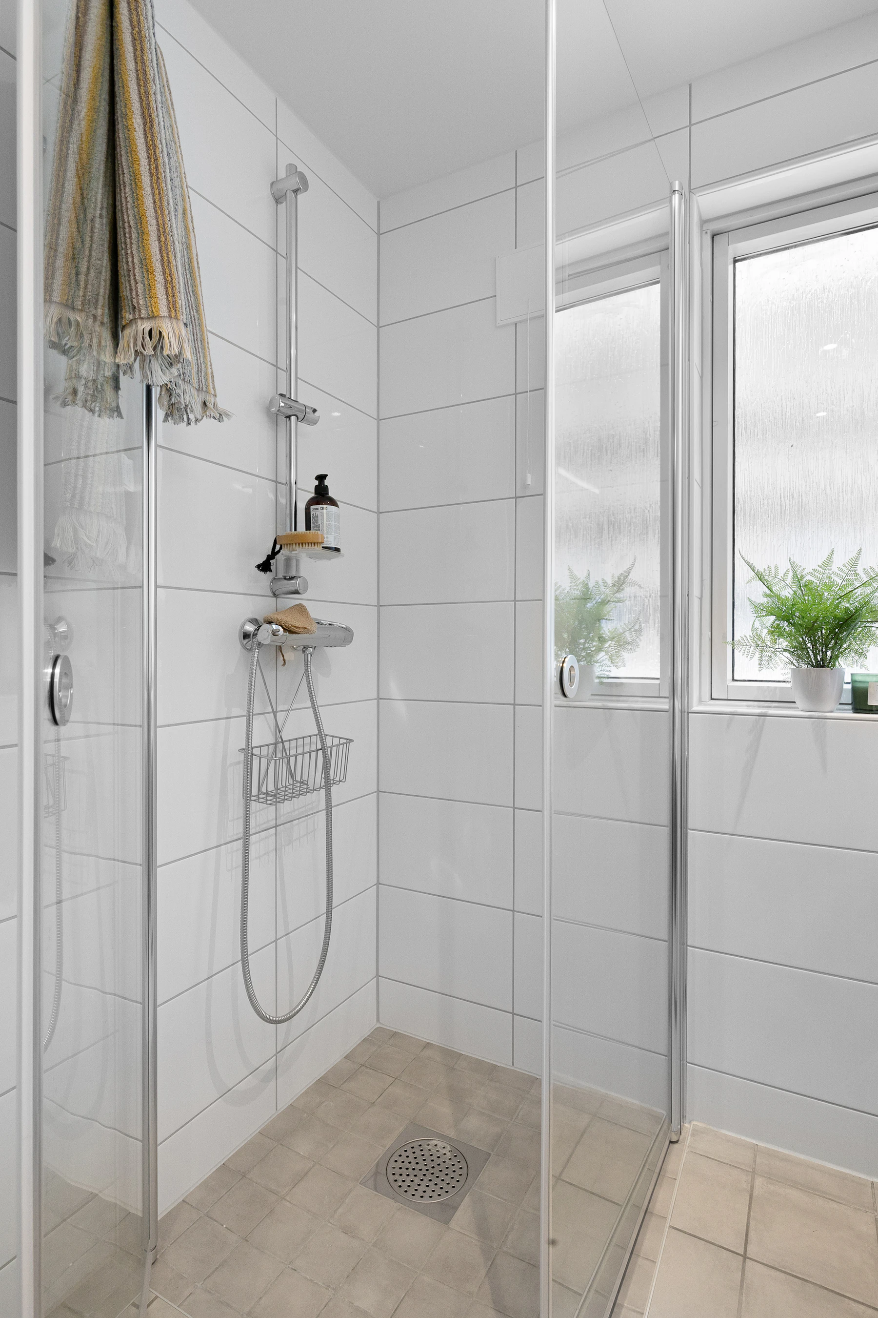 Walk-in shower with white tiled walls, glass doors, chrome rail shower, handheld fittings, wire basket, floor drain and frosted windows.