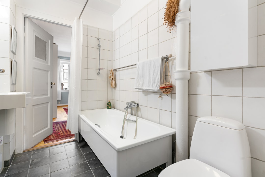 Simple renter bathroom with white wall tiles, white bathtub with shower curtain, wall-mounted towel rack, and a glimpse of a colorful hallway rug