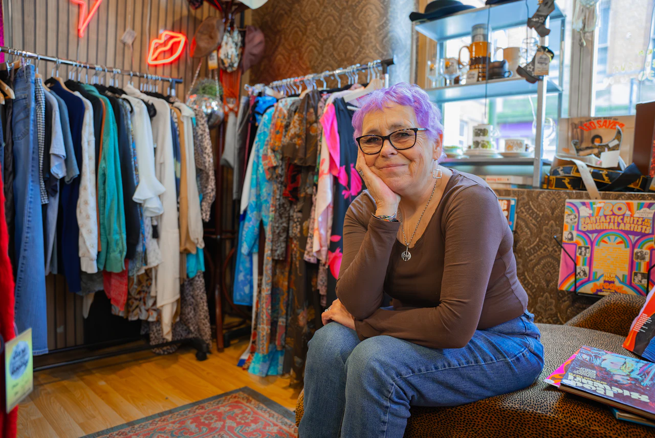 Woman with lilac hair sits smiling among rails of colourful clothes in a vintage shop.
