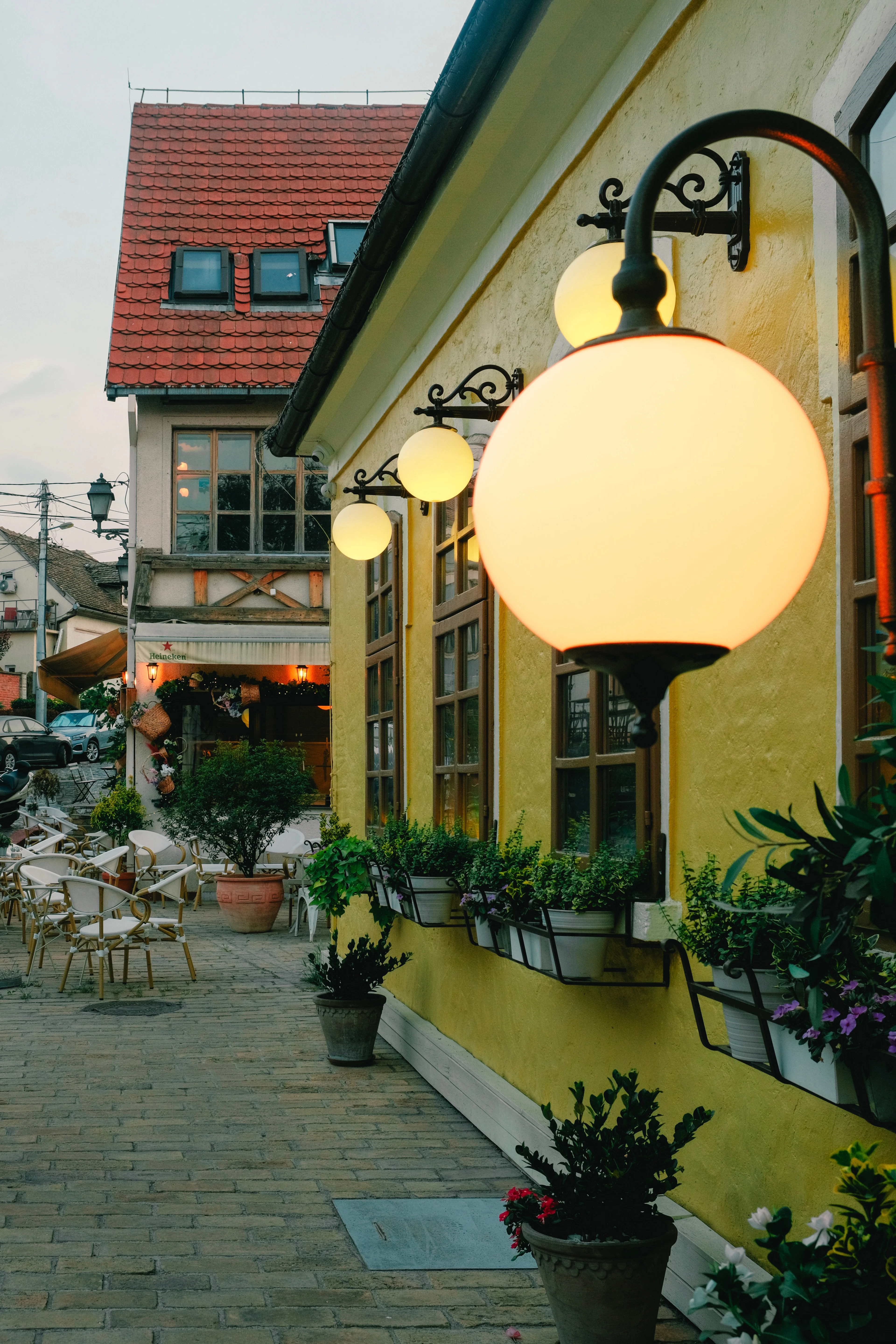Globe lamps glowing at dusk along the ochre-yellow wall of a Zemun tavern, window boxes of herbs and flowers below them and empty café chairs on the paving, with a steep red-tiled roof opposite.
