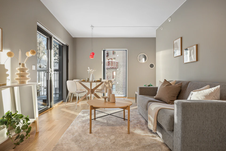 The image depicts a modern living room with light beige walls and a gray carpet, creating a warm and inviting atmosphere. A large window on the left wall allows natural light to flood into the space, illuminating the room. In the center of the room is a wooden coffee table surrounded by two white chairs, suggesting a cozy dining or social area.

On one side of the room, there's a gray sofa with a brown throw blanket draped over it, providing additional comfort and style. A red vase adds a pop of color to the space. The room also features a large window that offers a view of trees outside, further enhancing the natural ambiance.