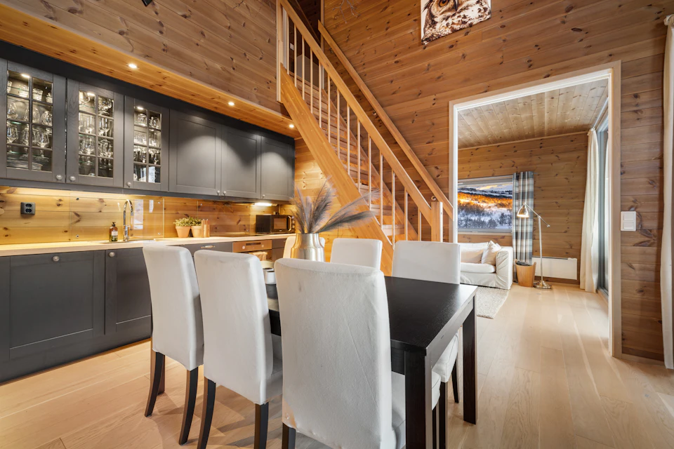 The image depicts a modern kitchen and dining area with a large wooden wall serving as the backdrop for the space. The kitchen features black cabinets, a white countertop, and a sink on one side. A staircase leads to another room, adding depth to the layout. In the center of the room is a table surrounded by six chairs, suggesting an inviting atmosphere for dining or socializing.

The dining area includes a large wooden table with four chairs surrounding it, positioned near a window that offers a view of the outdoors. A vase and potted plant are placed on the table, adding a touch of greenery to the space. The room is illuminated by recessed lighting, creating a warm and welcoming ambiance.