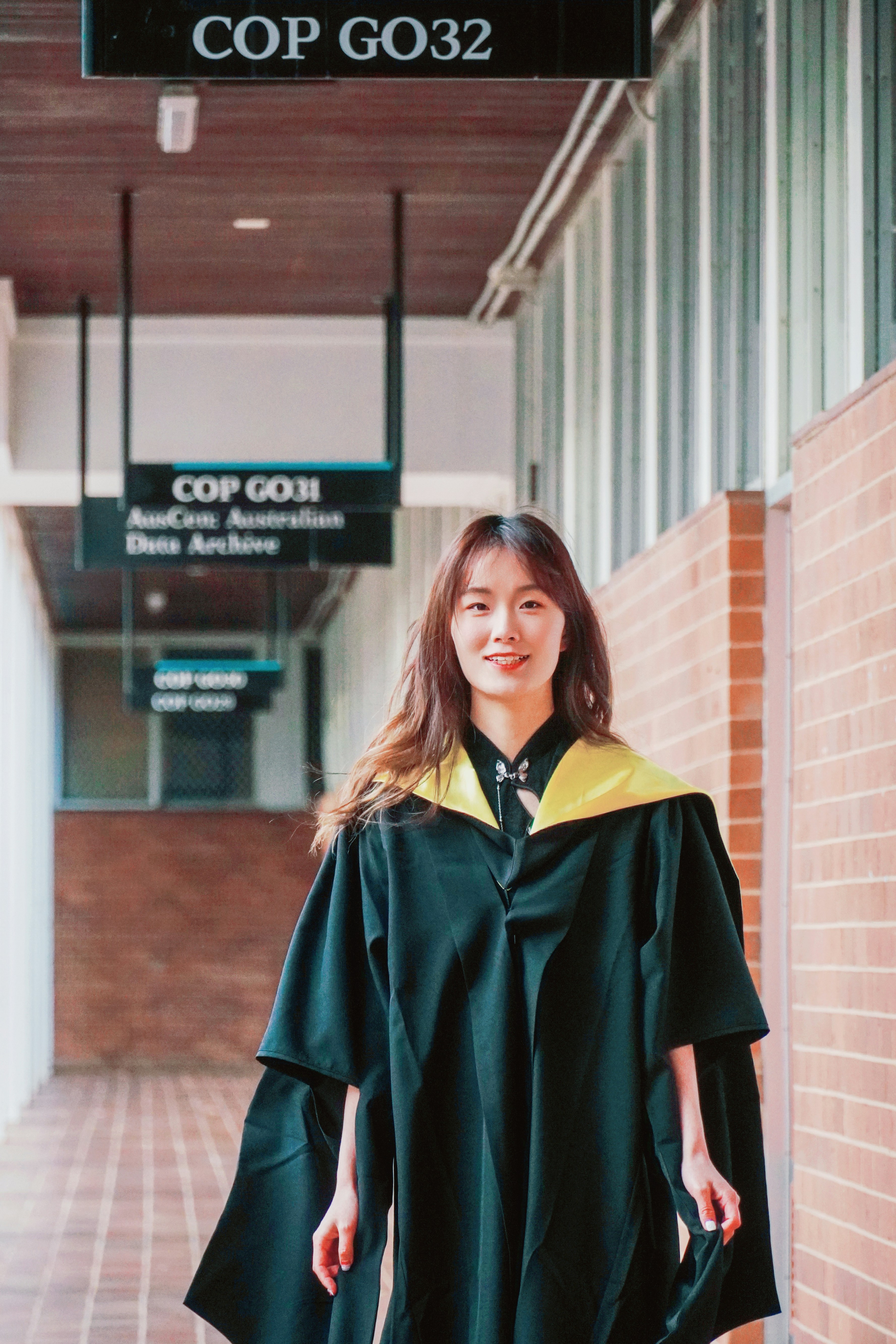 A graduate in a black gown with yellow accents walking down a corridor with signs overhead, with a joyful expression.
