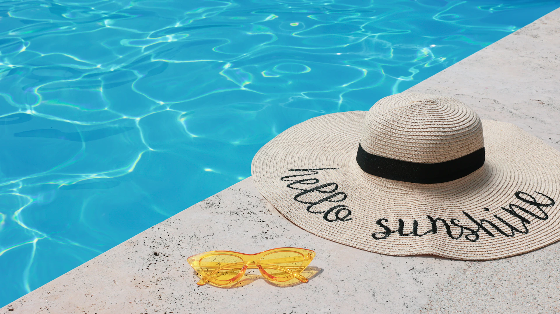 A hat, sunglasses, and a towel sitting on the edge of a swimming pool