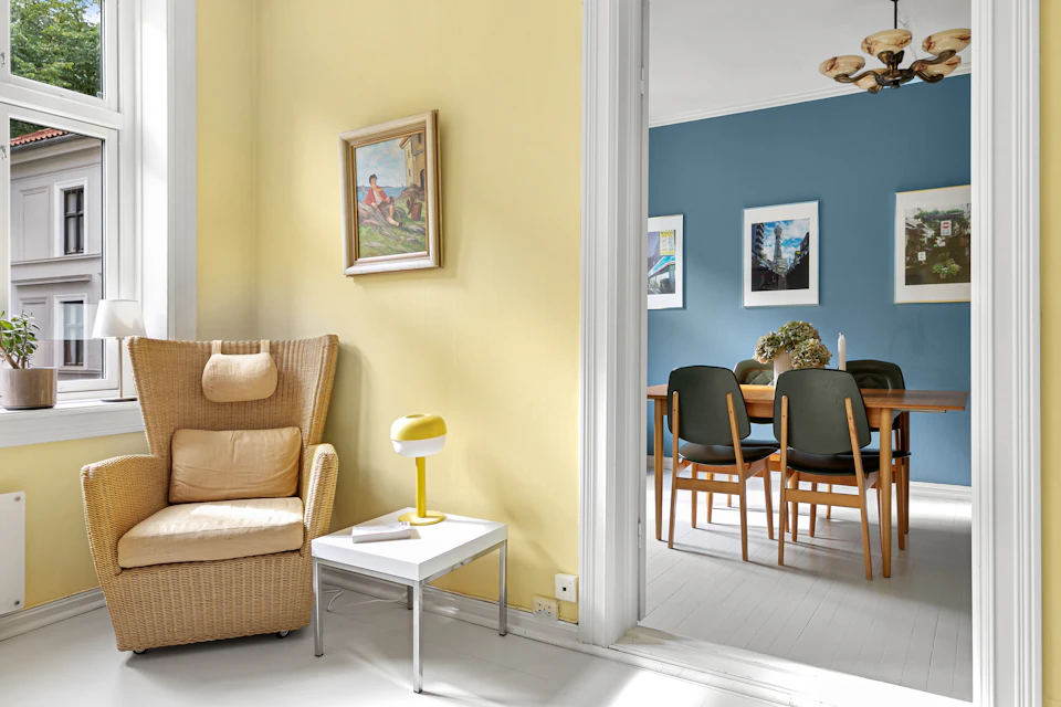 The image depicts a cozy living room with a large window on the left wall, allowing natural light to flood the space. The room features a beige armchair and a wooden coffee table, both adorned with a single yellow lamp that casts a warm glow throughout the room. A white door is visible in the background, leading to another part of the house. On the right side of the image, there's a dining area with a wooden table surrounded by four chairs, two on each side and one at the head of the table. The walls are painted in a light blue color, providing a calming backdrop for the room.
