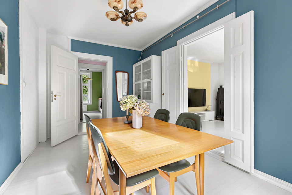 The image depicts a modern dining room with blue walls and white trim, creating a serene and elegant atmosphere. The centerpiece is a wooden table surrounded by four chairs, each paired with a matching chair on either side. A vase filled with flowers sits on the table, adding a touch of natural beauty to the space. In one corner of the room, there's an open door leading to another part of the house, suggesting that this dining area is part of a larger living space or a multi-functional home.
