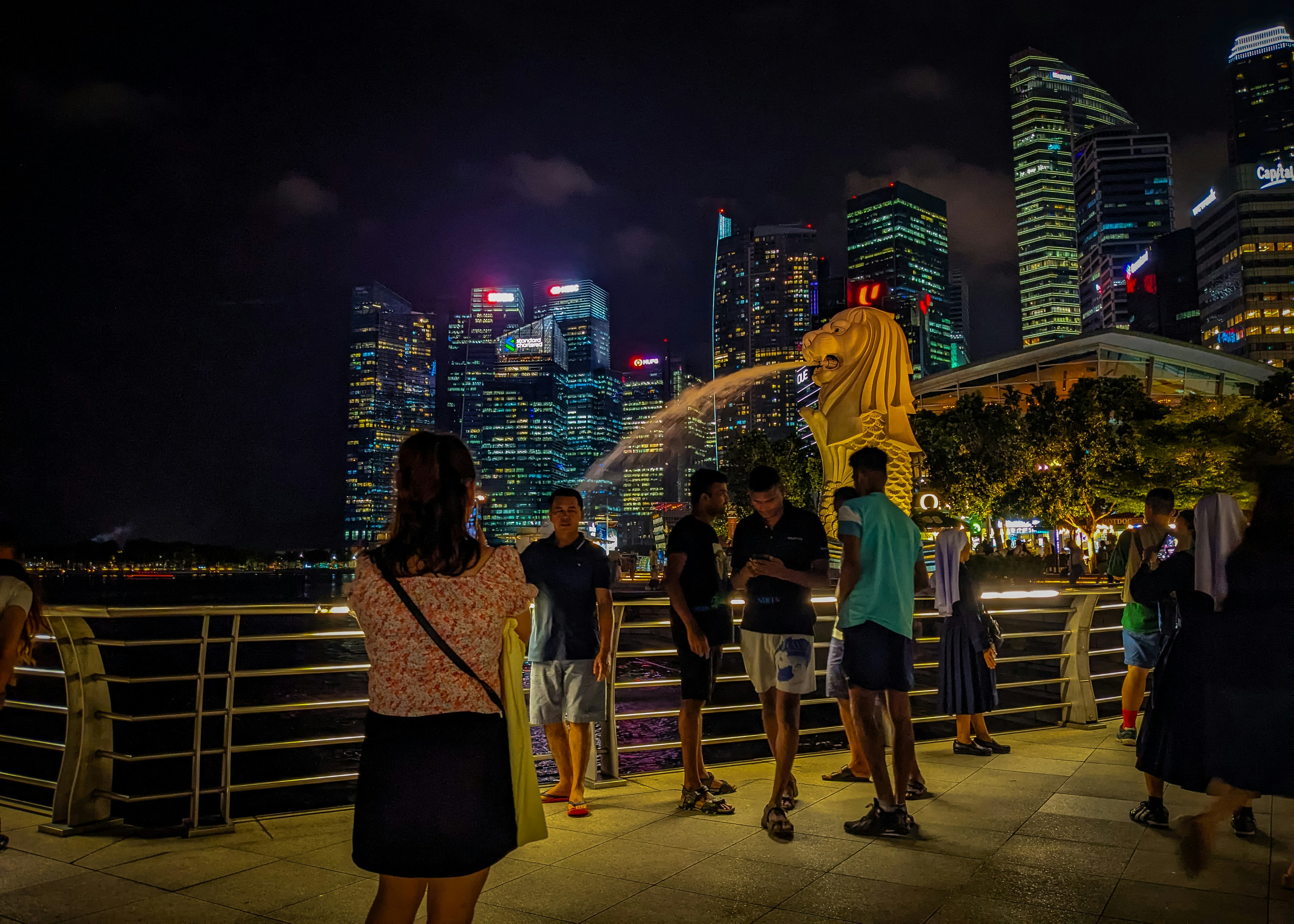 A nighttime shot of the iconic Merlion statue in Singapore, beautifully illuminated against the dark sky. Tourists gather around, capturing memories with their cameras as the Merlion's water stream glistens in the artificial lights. The vibrant city lights in the background add to the dynamic and lively atmosphere, showcasing a blend of cultural significance and modern allure.
