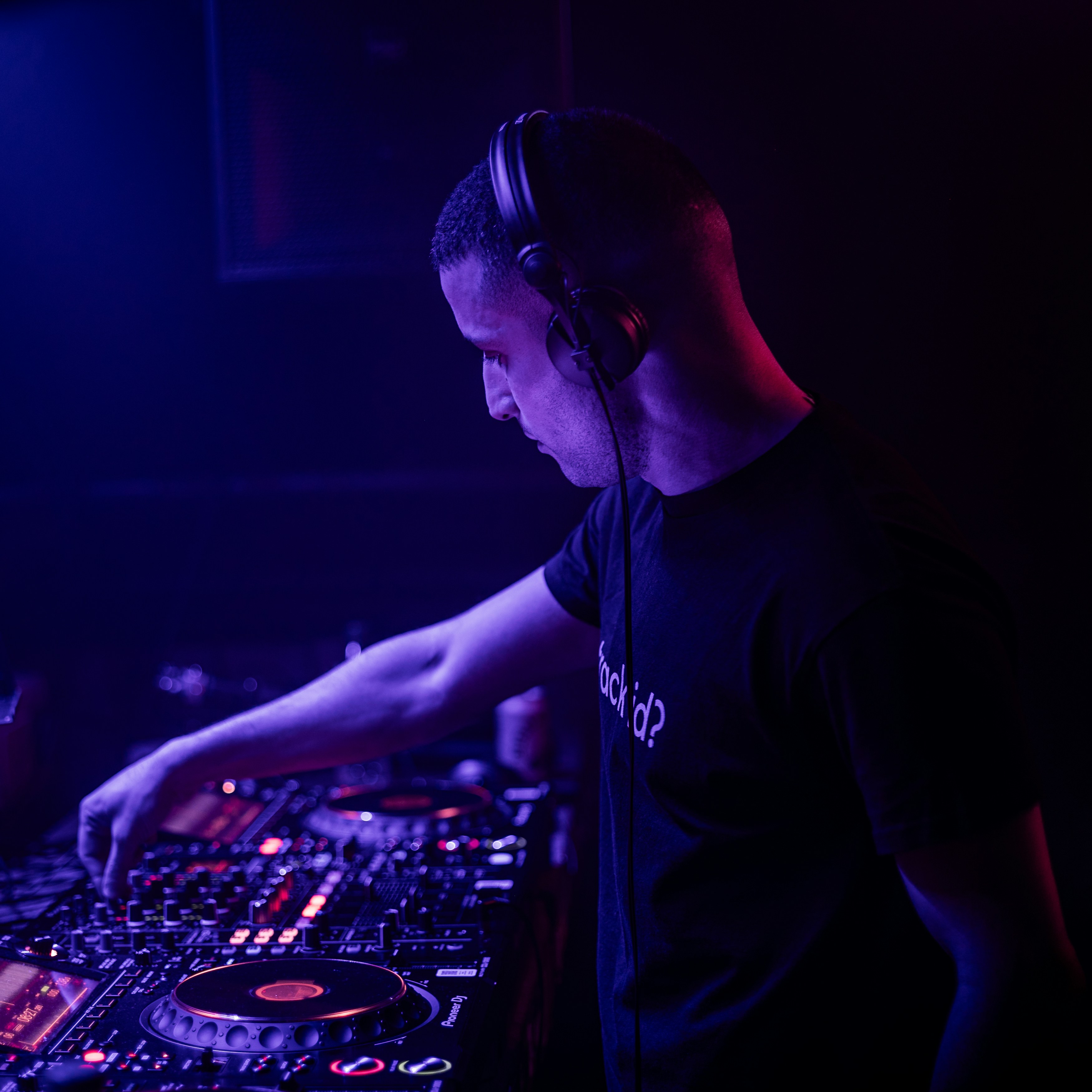 Professional DJ at Pioneer CDJ setup in a dark venue focused on mixing, blue and purple lighting illuminating equipment, confident professional atmosphere