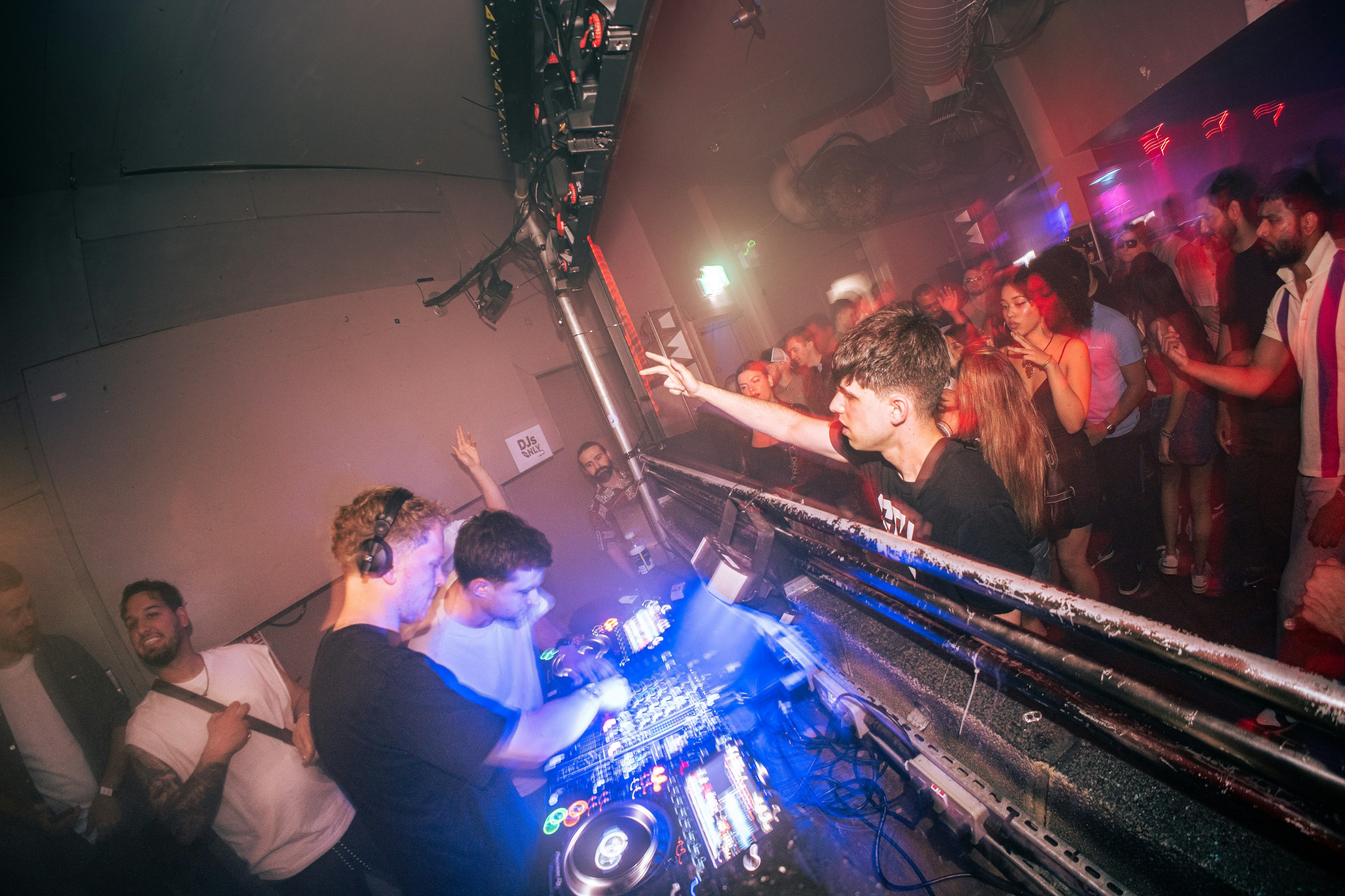 DJ performing at a packed nightclub with coloured lighting beams, dark atmospheric venue, crowd in silhouette