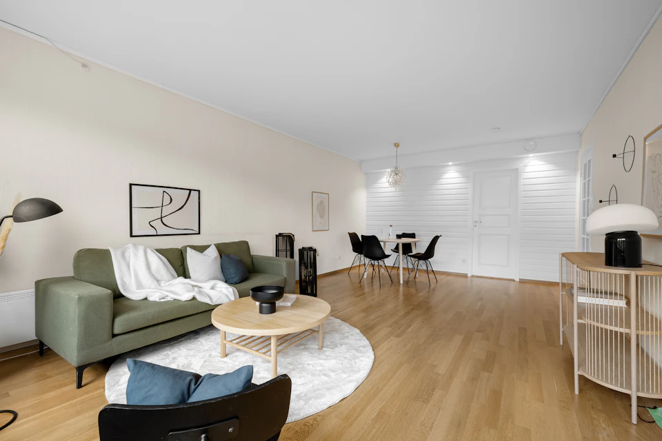 The image depicts a modern living room with a large window that allows natural light to flood the space, illuminating the white walls and wooden floor. The room features a green sofa adorned with blue throw pillows, positioned against one wall. Adjacent to it is a black coffee table hosting a vase of flowers. In front of the sofa, there are two chairs - one black and one gray. A round rug lies in the center of the room, adding warmth and texture to the space. On the right side of the image, a white dining table with four chairs stands ready for meals or gatherings. The walls are painted white, creating a bright and airy atmosphere, while the wooden floor adds a touch of rustic charm.