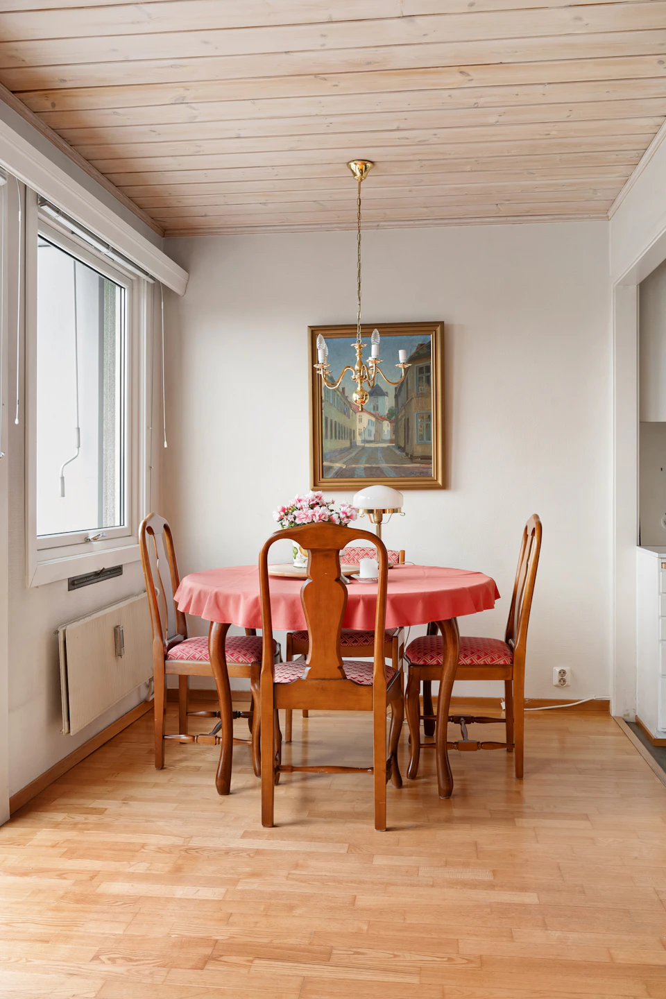 The image depicts a cozy dining room with a wooden floor and white walls, creating an inviting atmosphere. A round table is set for four on the right side of the room, adorned with two chairs each. The table is draped in a vibrant red tablecloth, adding a pop of color to the scene. Above the table hangs a painting that adds an artistic touch to the space. On one side of the room, there's a window offering natural light and a view of trees outside. A chandelier hangs from the ceiling, casting a warm glow over the dining area. The walls are painted white, which complements the wooden floor and enhances the overall brightness of the room.
