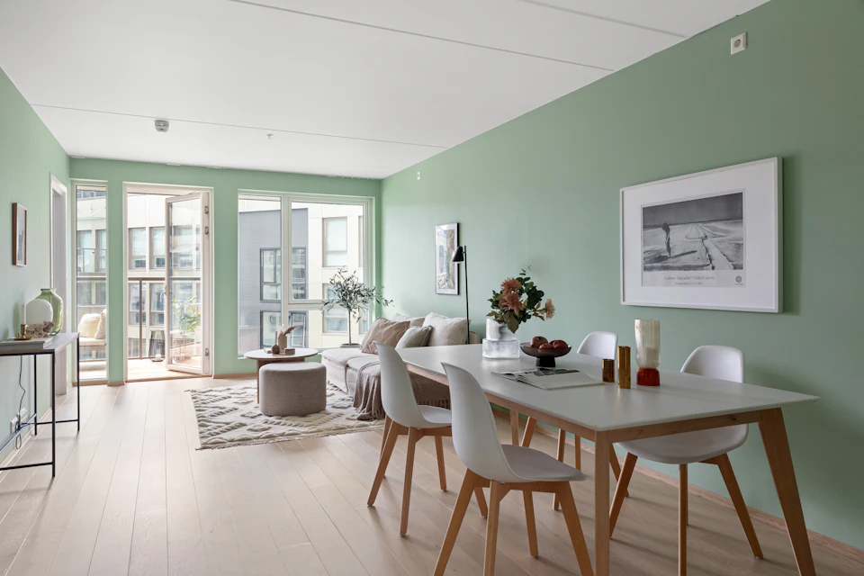 The image depicts a modern, open-concept living room and dining area with light green walls that reflect off the white ceiling. The room features a large window on the left wall, allowing natural light to flood the space. A gray rug covers the floor in front of a wooden coffee table, which holds a vase filled with flowers.

The dining area is furnished with four chairs surrounding a white table draped with a white tablecloth and set against the green walls. The room also includes a couch situated on the left side, creating an inviting atmosphere for relaxation or socializing. A few potted plants are scattered throughout the space, adding a touch of greenery to the room.