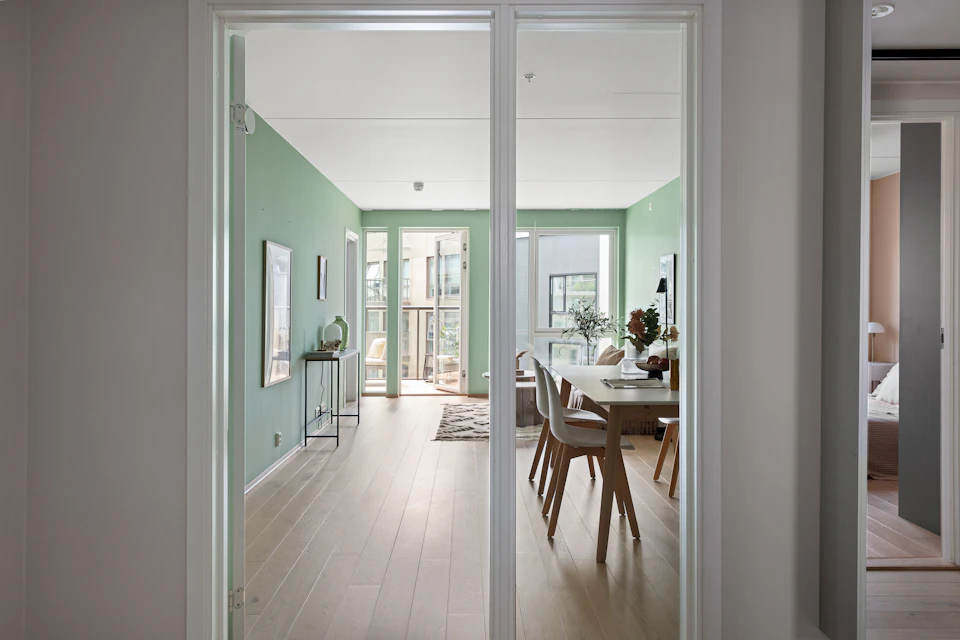 The image shows a modern, open-concept living room with a large window that allows natural light to flood the space. The room features a white color scheme and is furnished with a couch on one side and a dining table surrounded by chairs on another side. A green door can be seen in the background, adding a pop of color to the otherwise neutral palette.