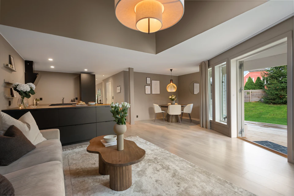 The image depicts a modern living room with a large window that offers a view of a backyard, allowing natural light to flood the space. The room features a gray sofa and a coffee table adorned with a vase filled with white flowers, creating a serene atmosphere. A dining table is positioned in the center of the room, surrounded by four chairs. In one corner, there's an open door leading to another part of the house, suggesting an open floor plan that connects different areas seamlessly. The walls are painted in a light beige color, and the ceiling has recessed lighting fixtures.